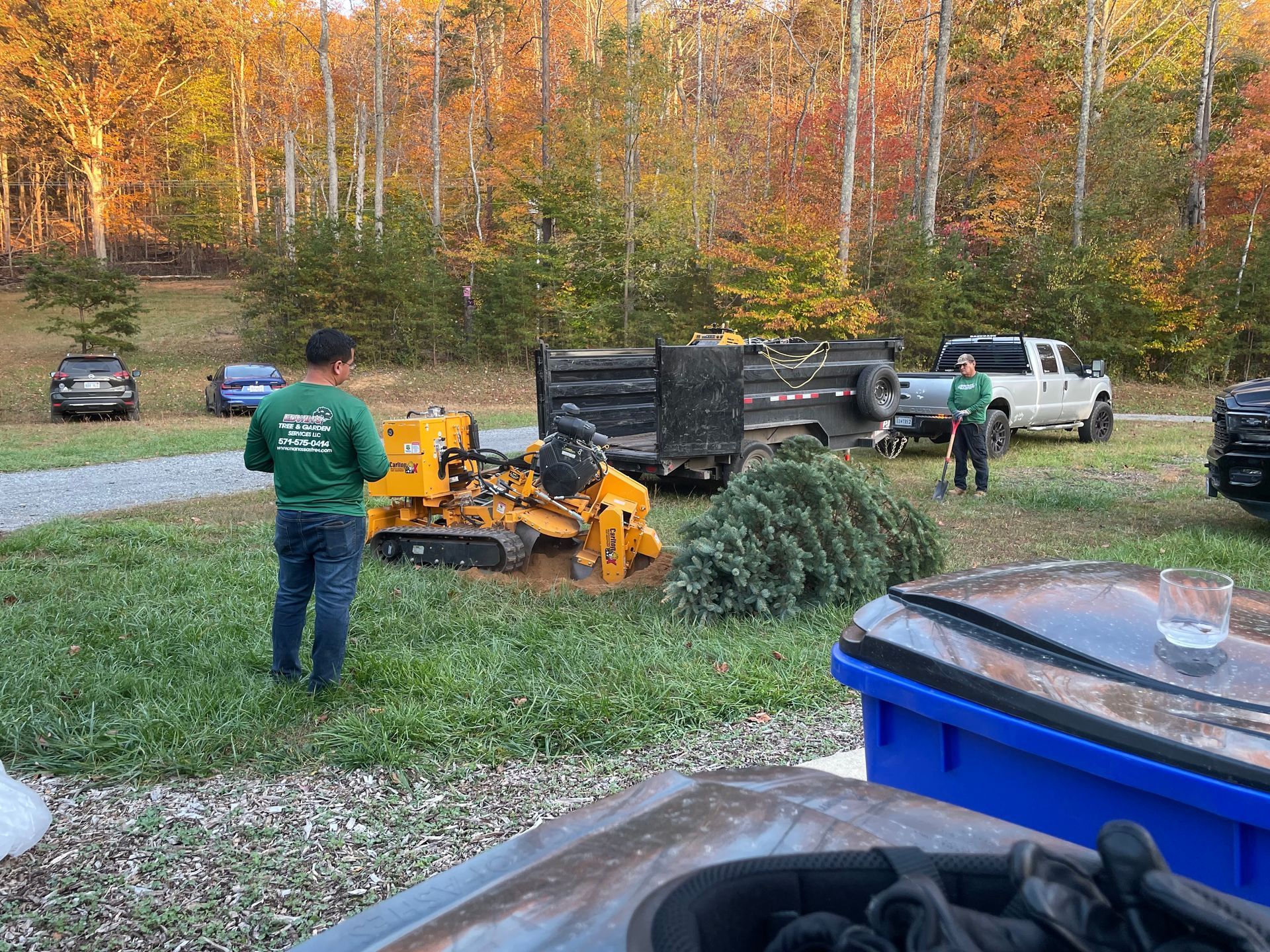 Two people grinding a tree stump outdoors, with equipment, a truck, and fall foliage.