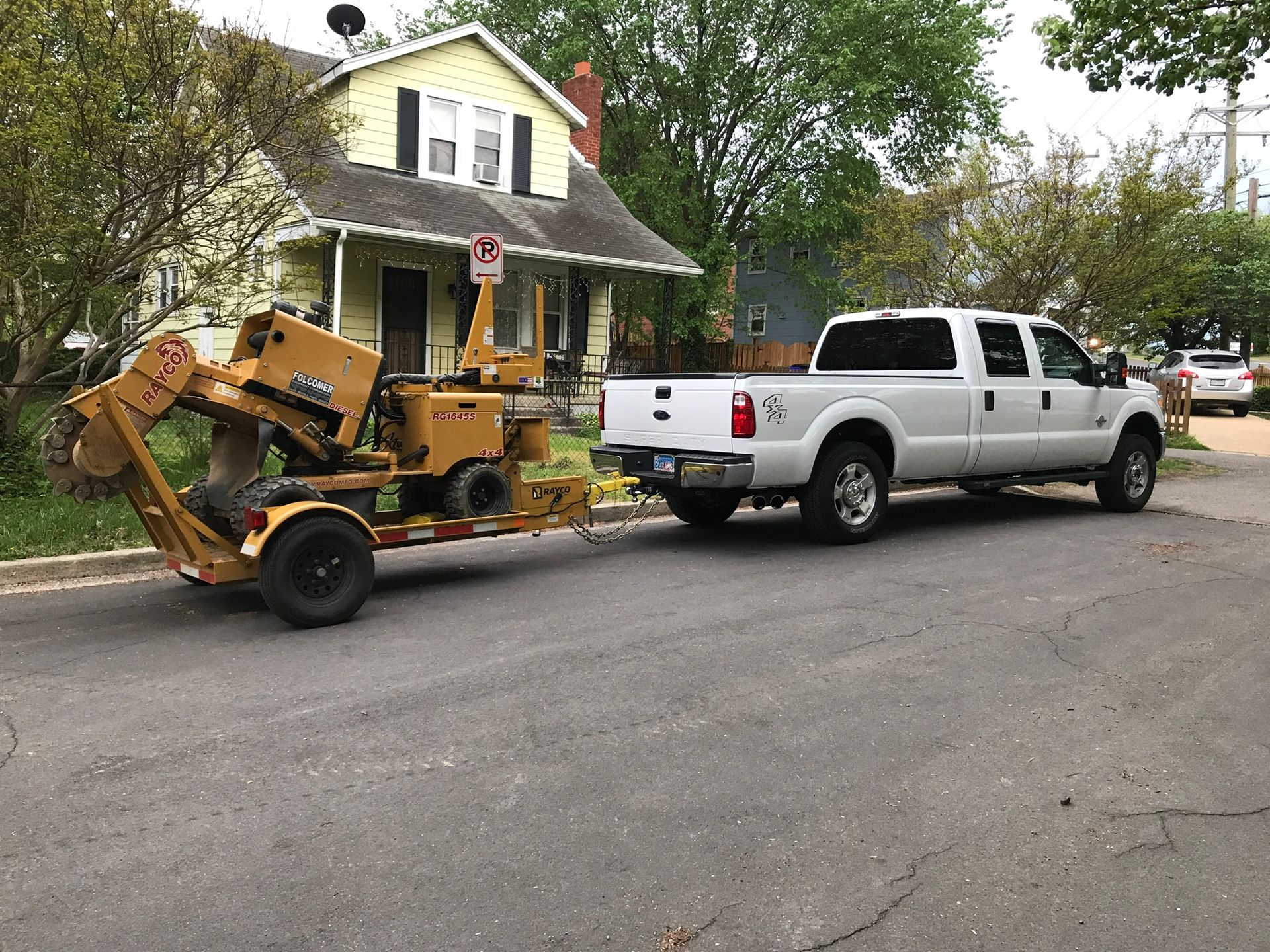 White truck towing a yellow stump grinder on a residential street.