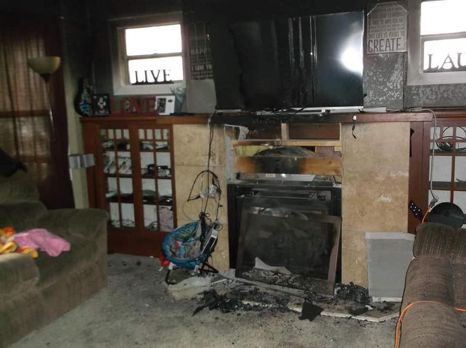 Living room interior after a fire. Charred fireplace, damaged furniture, and debris are visible.