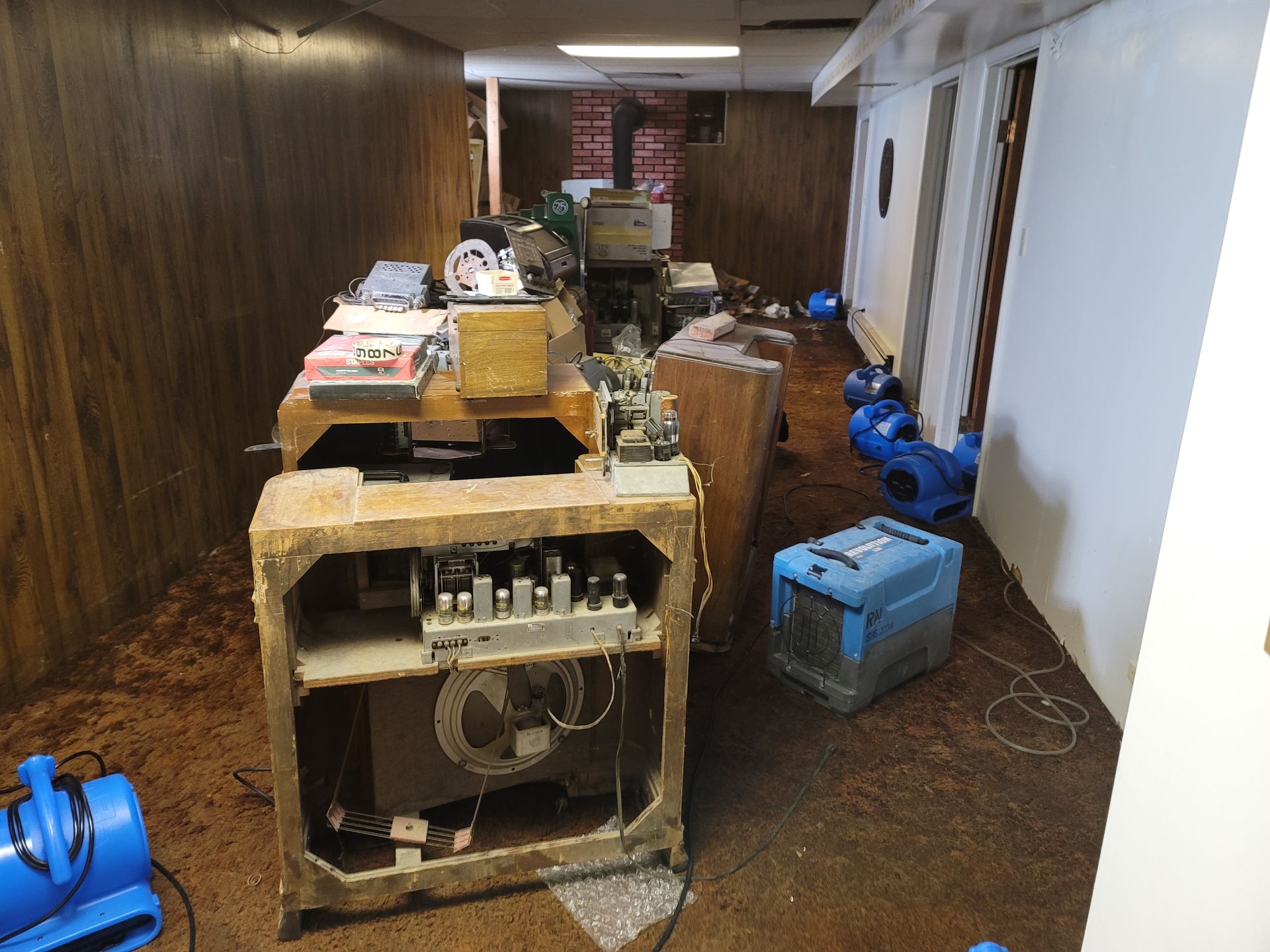 Flooded basement with vintage radio equipment, debris, and blue air movers.