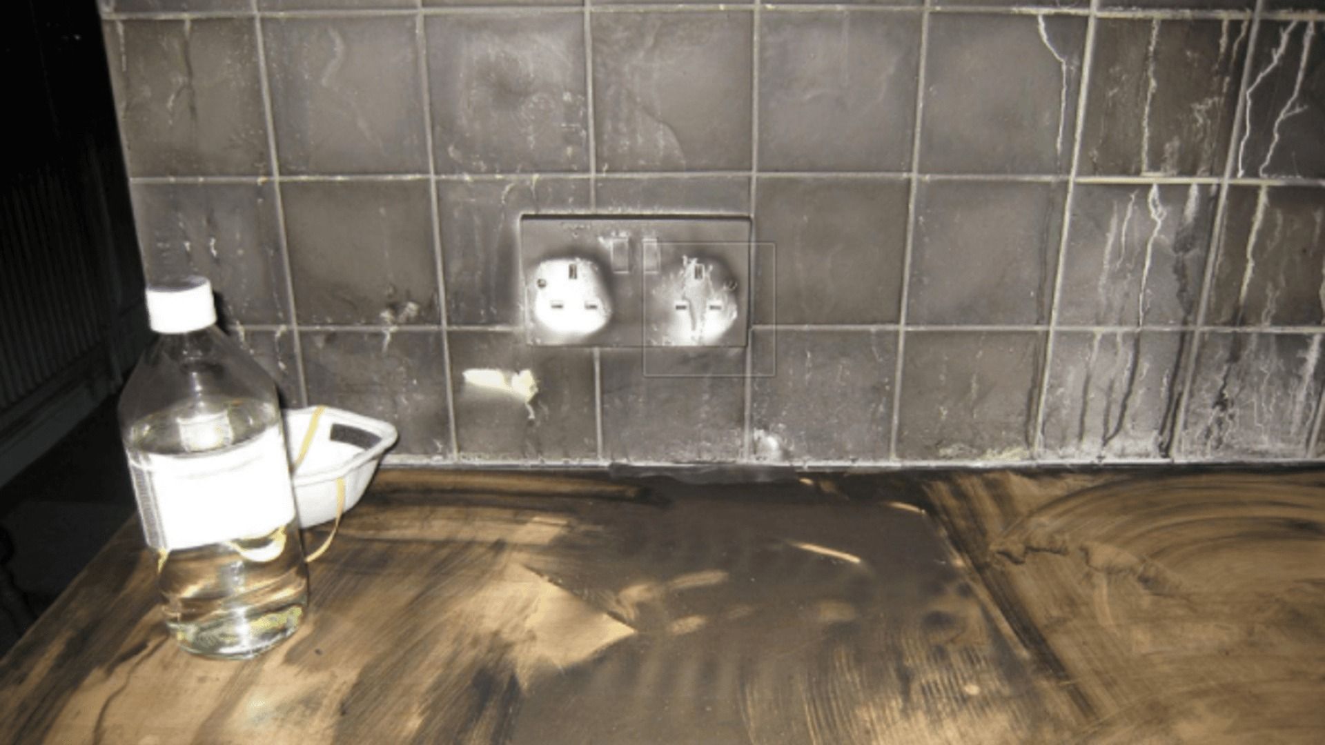 Charred kitchen wall with outlets, countertop, and bottle; evidence of a fire.