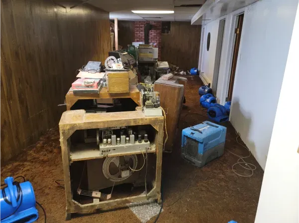 Flooded basement with damaged electronics and furniture; air movers are drying the room.