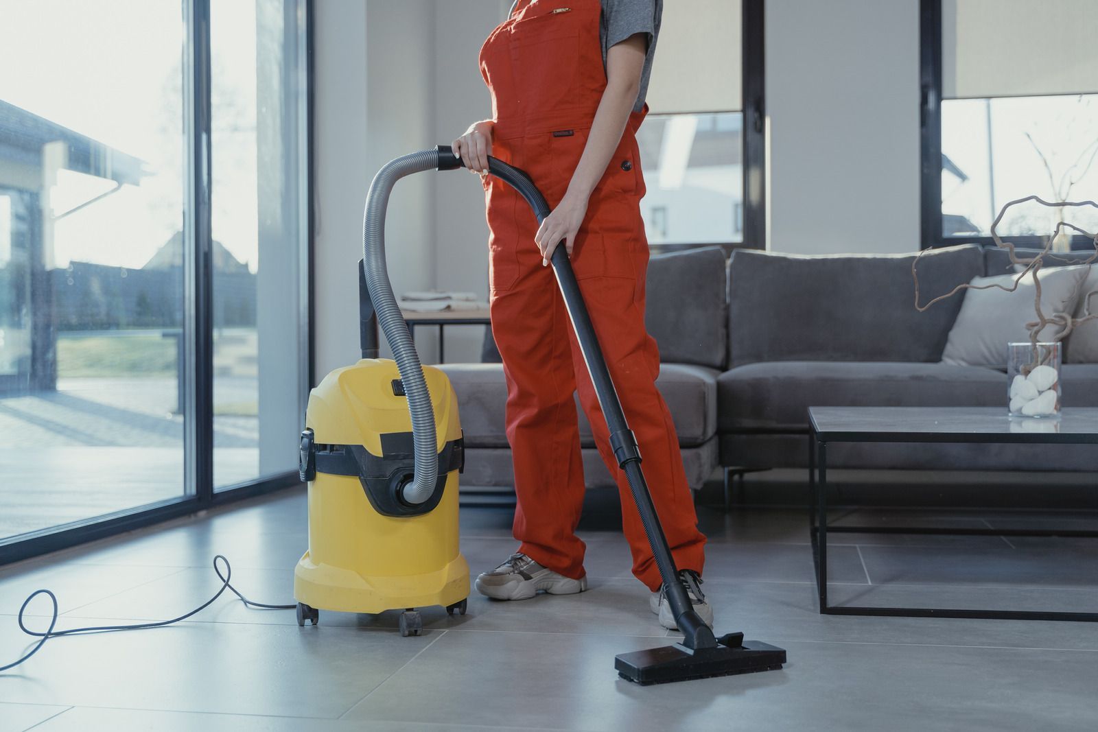 A person in red overalls vacuuming a light-filled living room with a yellow vacuum cleaner.