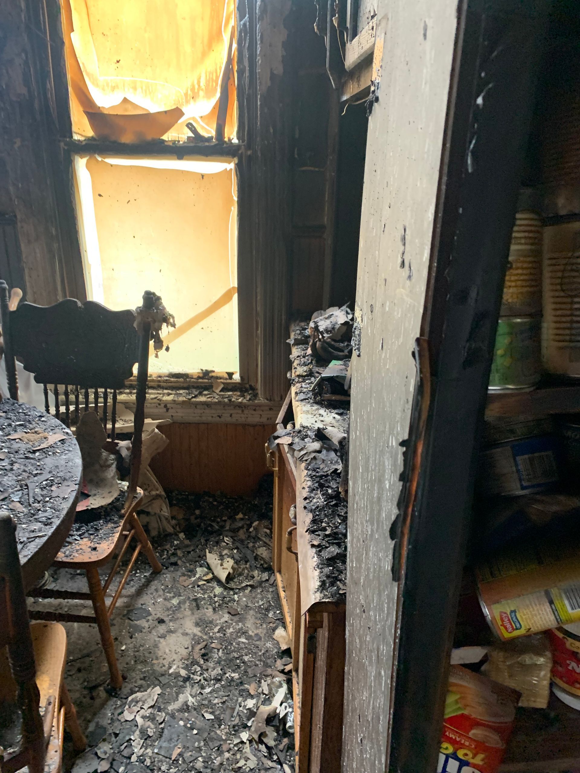 Interior view of a room heavily damaged by fire. Burnt furniture, debris, window with light visible.