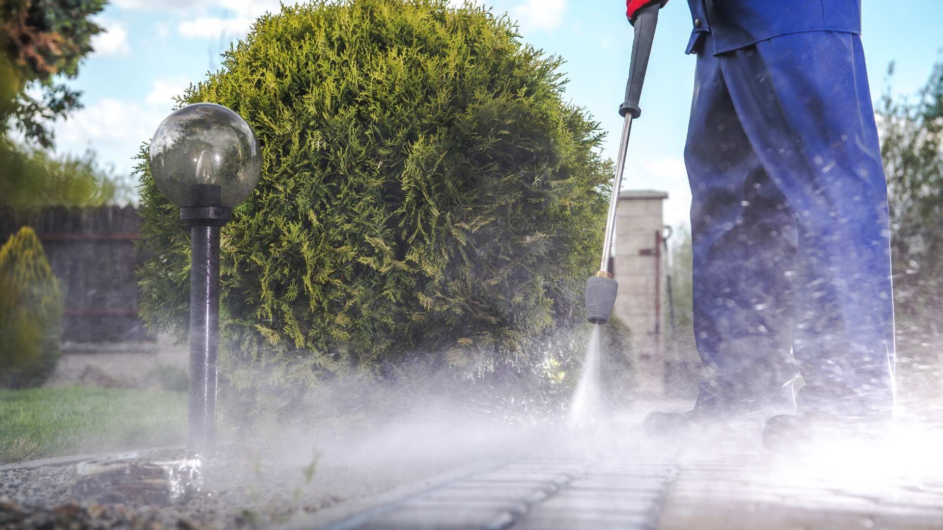 Person in blue overalls pressure washing a brick patio. Water spray and a garden.