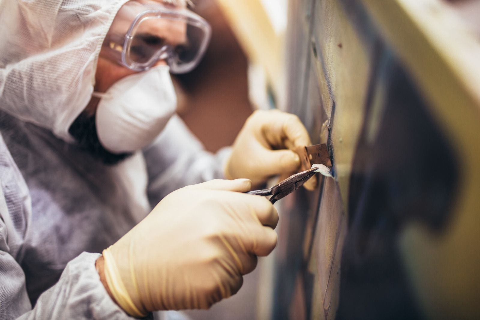 Person in protective gear scrapes peeling paint from a wall.