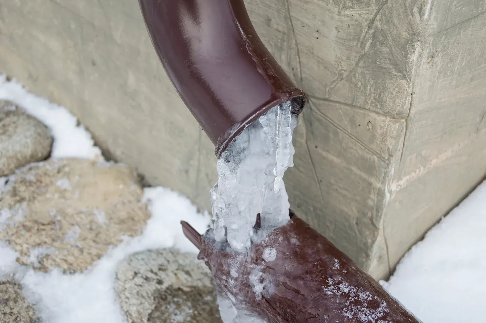 Icy, brown downspout on a building with ice buildup and snow on the ground.