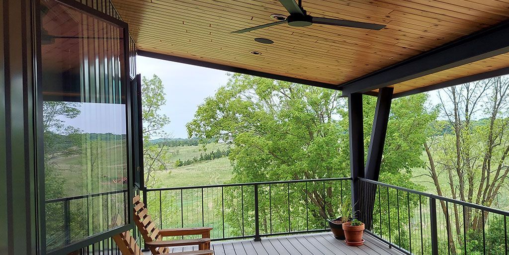 Balcony overlooking green trees and a distant hill, with a wooden chair, pots, and ceiling fan.