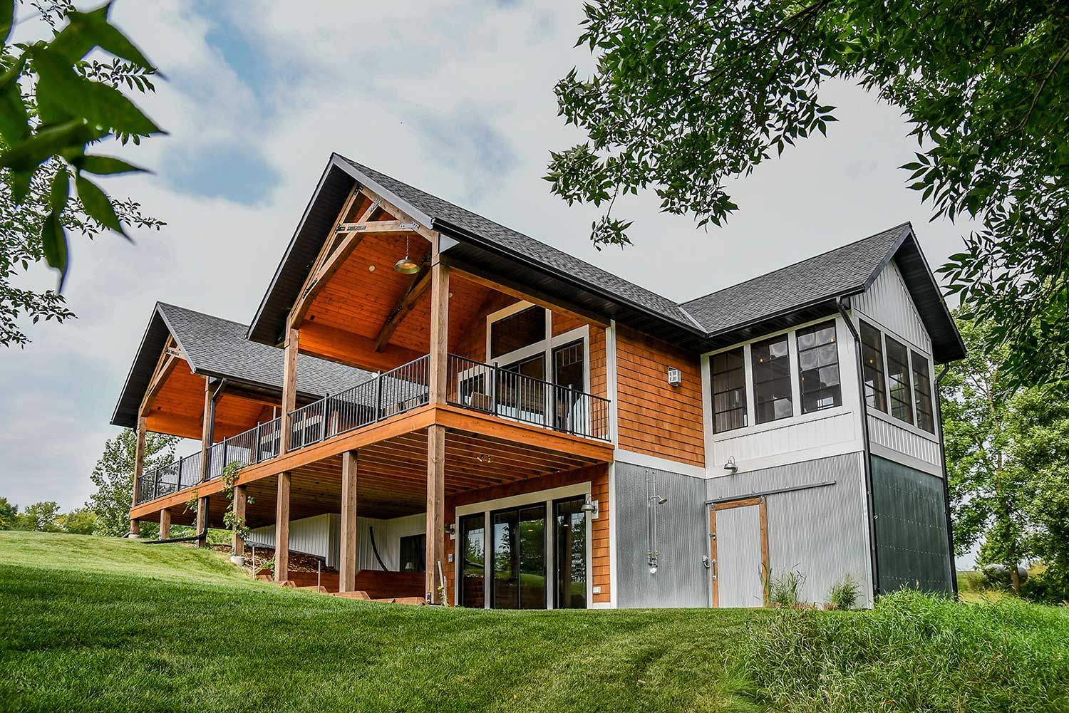 Two-story wood home with large deck, set on a grassy hill under a cloudy sky.