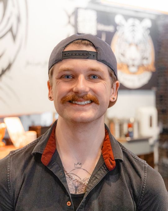 Man with a mustache and cap smiles, in front of the tigers den logo. He wears a gray shirt with red trim.
