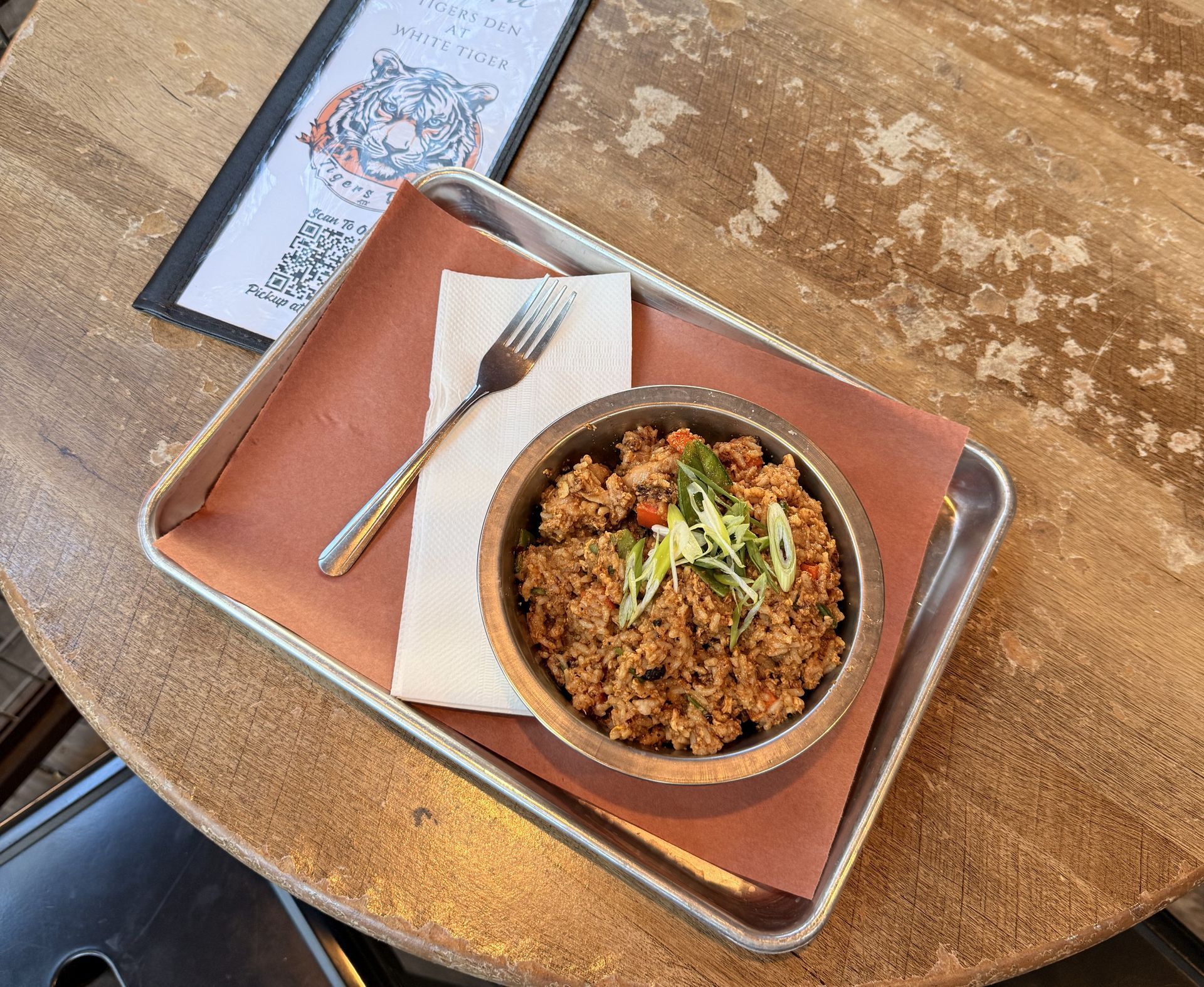 Metal bowl of spicy fried rice topped with scallions on a tray with a napkin and fork, placed on a worn wood table next to a Tigers Den menu.