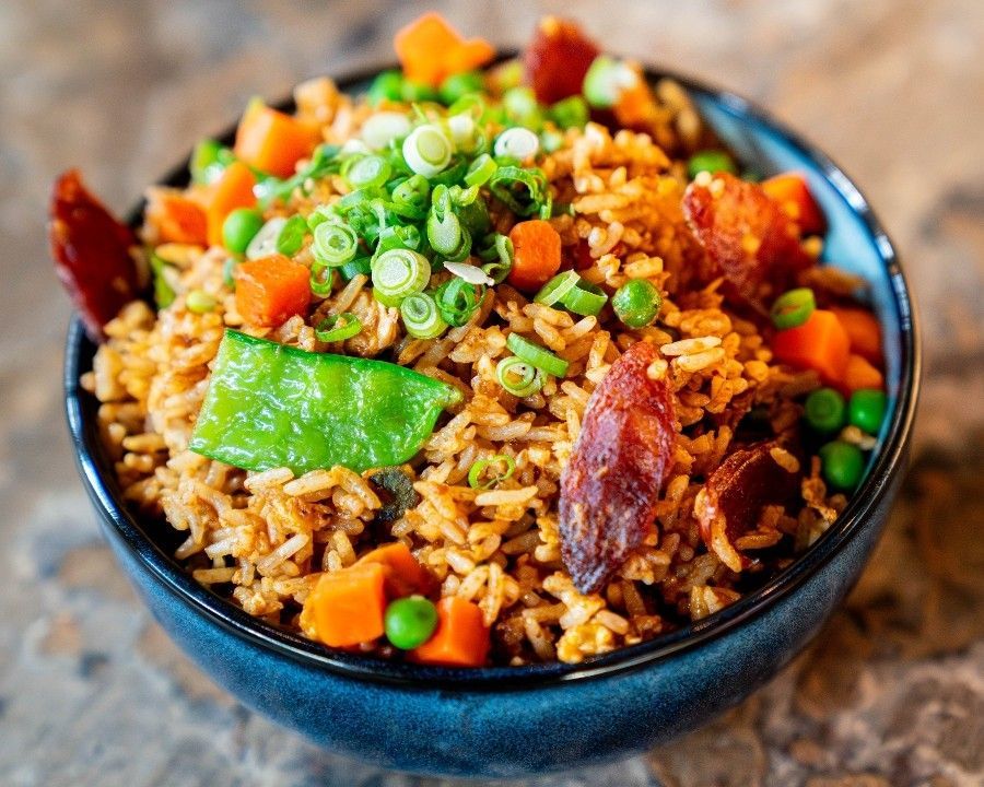 Colorful bowl of fried rice with green onions, snow peas, carrots, peas, and slices of Chinese sausage, served in a black bowl on a stone-textured surface at Tigers Den.