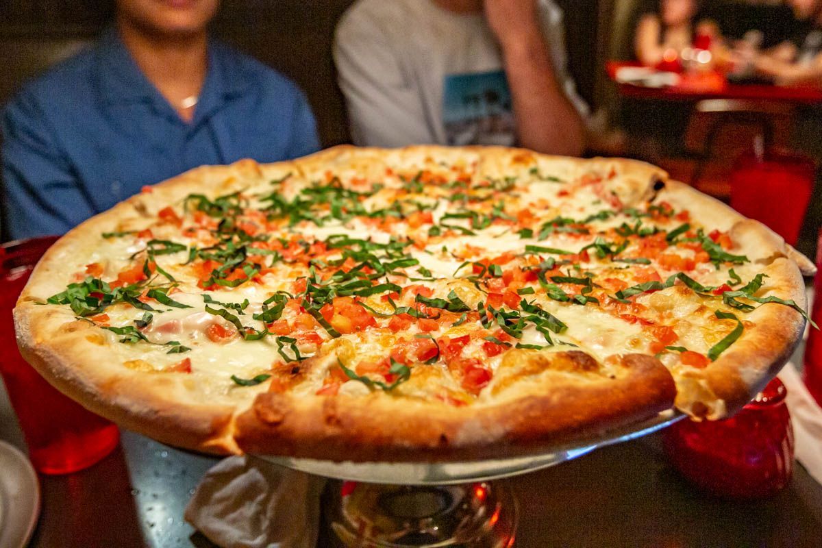 A freshly baked Margherita pizza from Home Slice Pizza in Austin, topped with melted mozzarella, diced tomatoes, and fresh basil on a golden, crispy crust, served on a raised pizza tray in a lively dining setting.