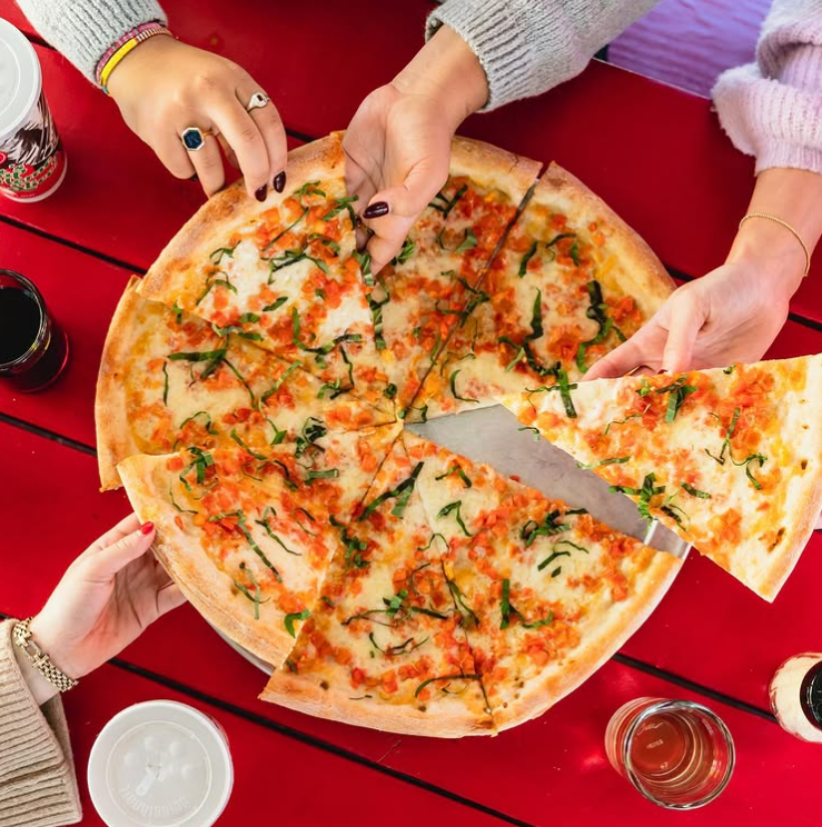 Hands reaching for slices of a large pizza topped with herbs and cheese on a bright red outdoor table, surrounded by drinks at Home Slice Pizza in Austin.