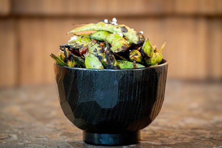 Black bowl filled with grilled edamame topped with sesame seeds and chili flakes on a wooden table.