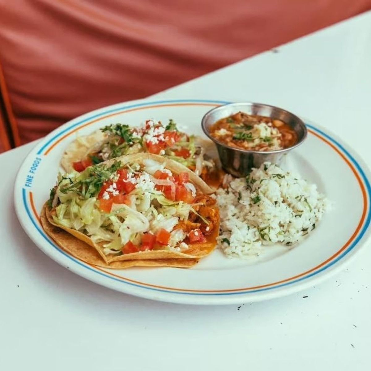 Three crispy tacos topped with lettuce, tomato, and cotija cheese served with cilantro rice and charro beans on a white diner-style plate at Joann&rsquo;s Fine Foods in Austin.