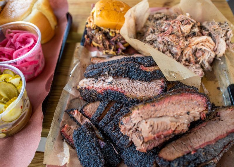 A tray of smoked Texas barbecue from La Barbecue in Austin, featuring thick-cut brisket, pulled pork, and a chopped brisket sandwich, served with pickles and onions.