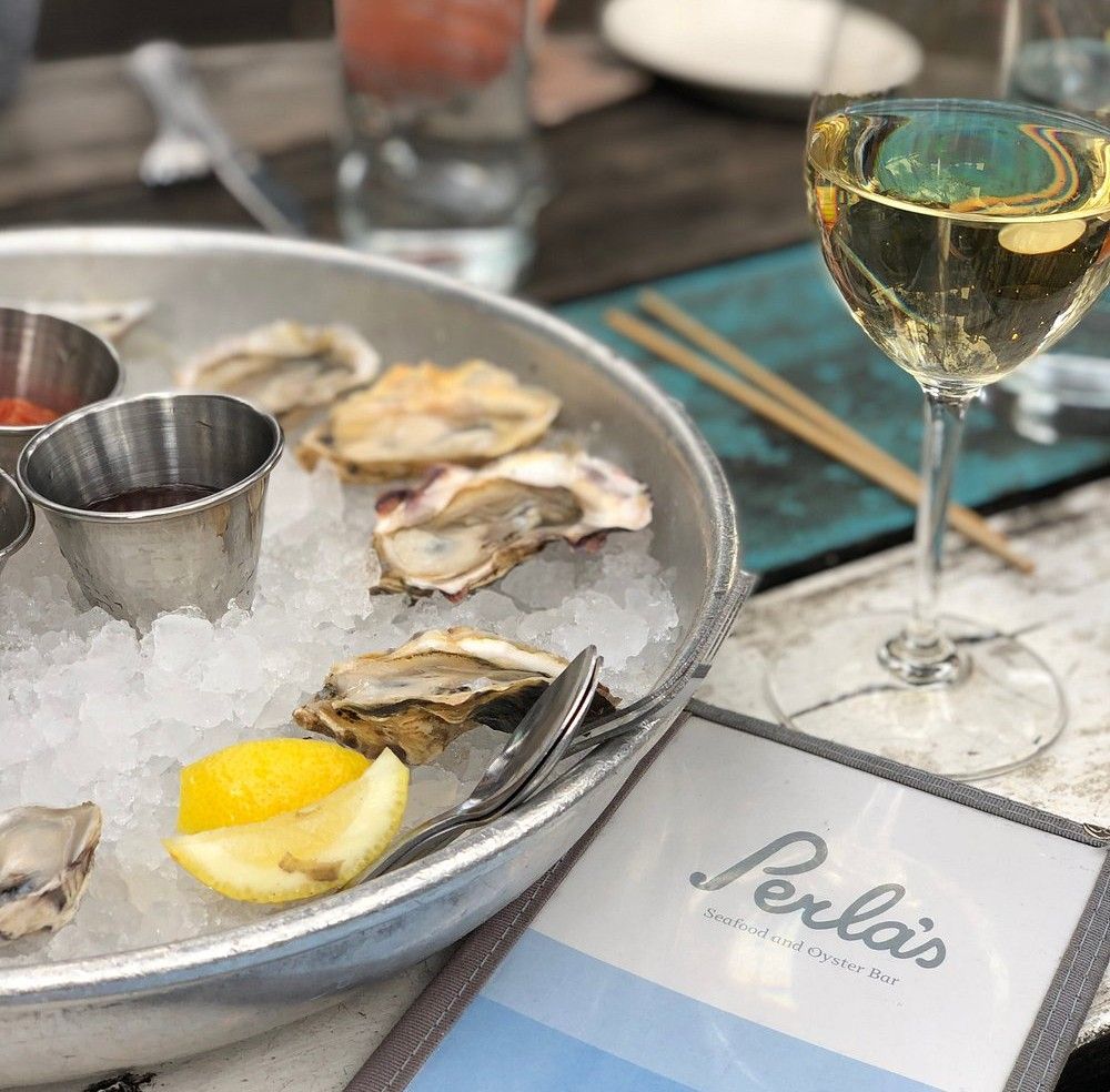 Close-up of a chilled oyster platter on ice with metal sauce cups and a lemon wedge, alongside a glass of white wine and a Perla’s menu at an outdoor table.