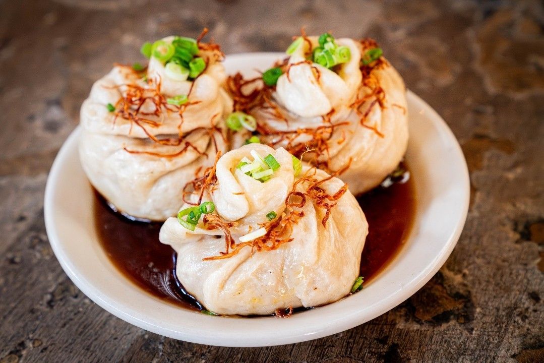 A plate of three pork-stuffed bao buns at Tiger’s Den, topped with crispy fried shallots and fresh green onions, sitting in a pool of rich, savory sauce.