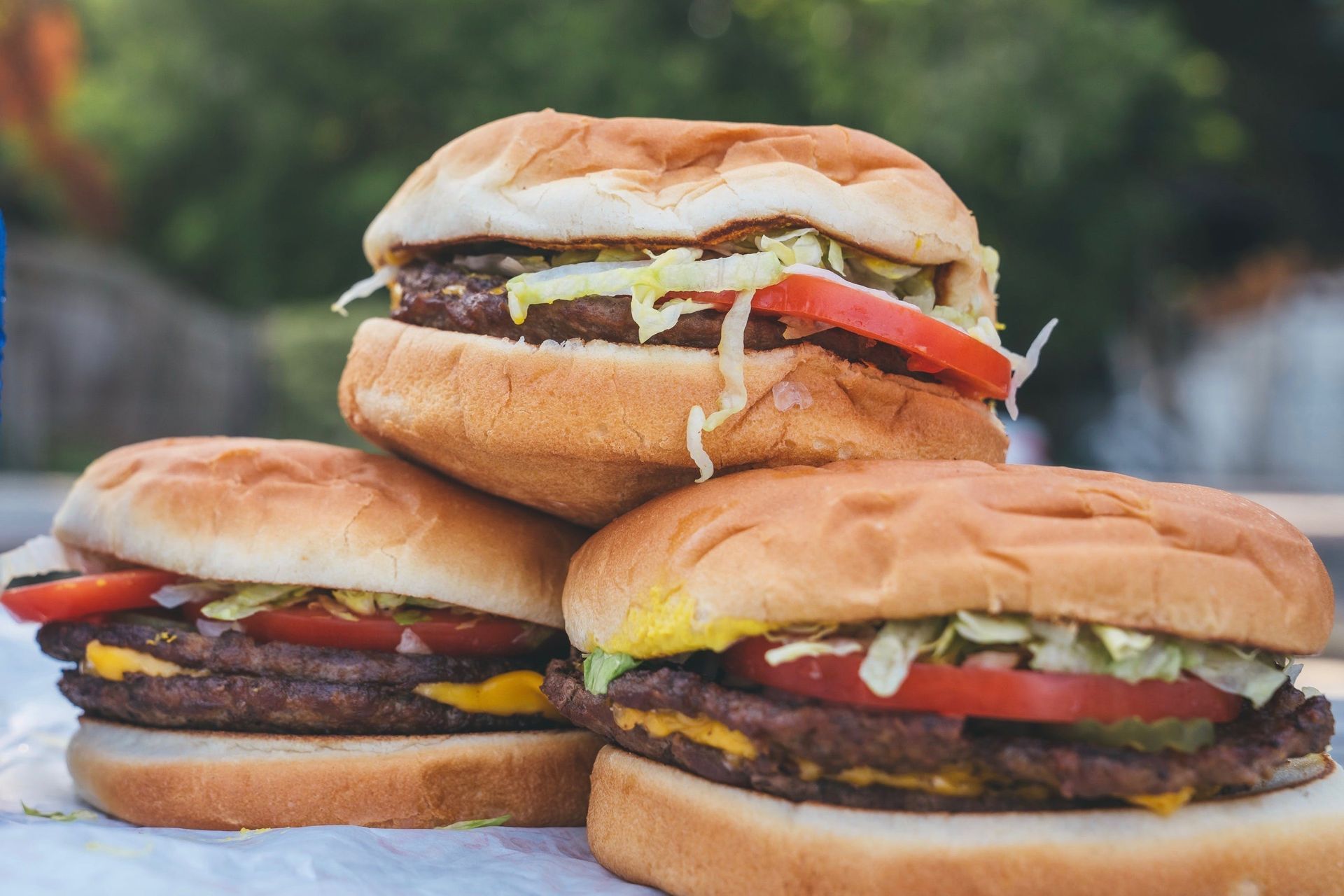 Three stacked cheeseburgers with lettuce, tomato, pickles, and mustard on a paper wrapper, with a soft focus green background, served outdoors at Sandy’s Hamburgers in Austin.