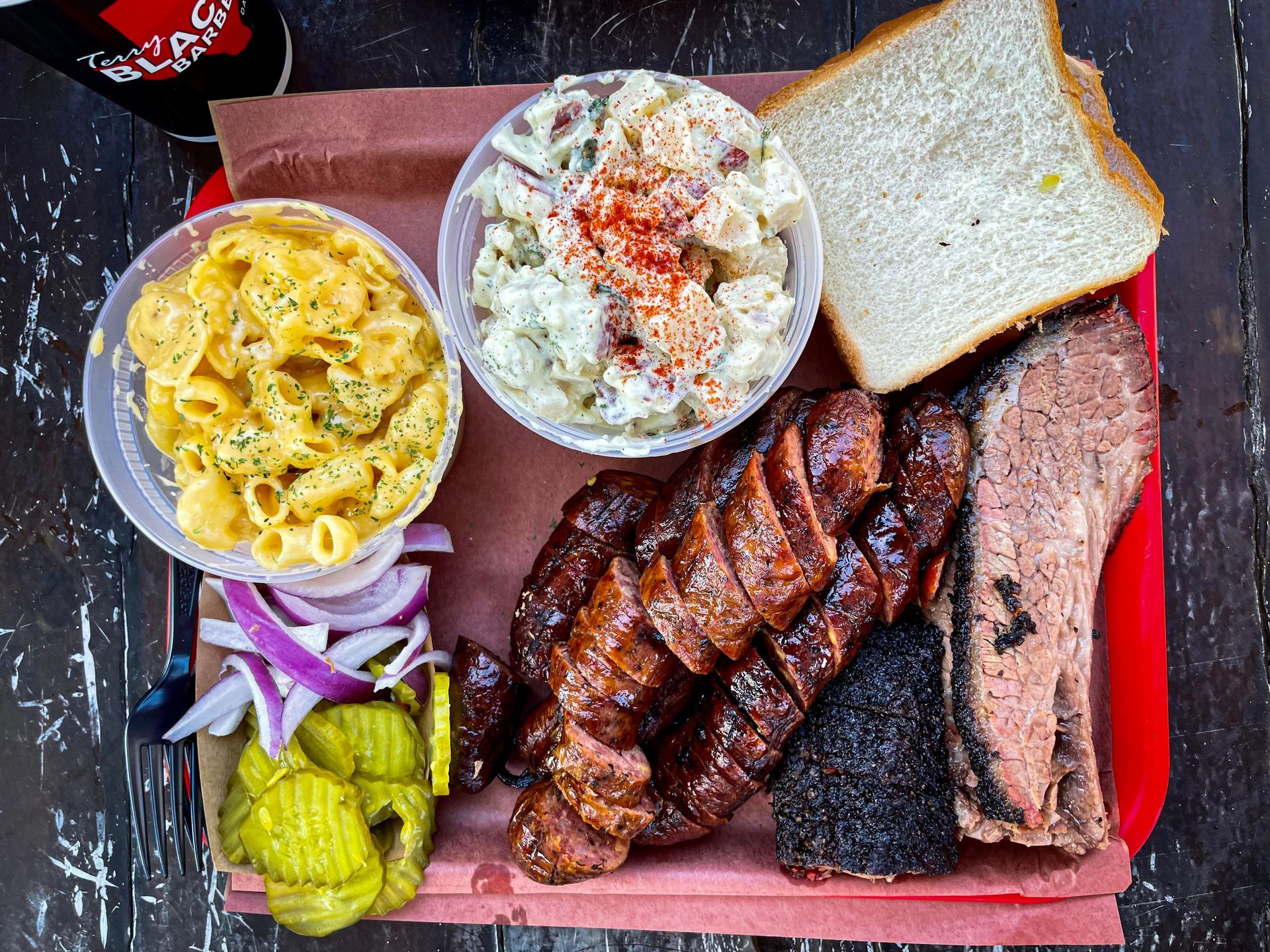 Barbecue tray with sliced brisket, sausage, mac and cheese, potato salad, pickles, onions, and white bread on a red tray with butcher paper, served at Terry Black’s Barbecue in Austin.