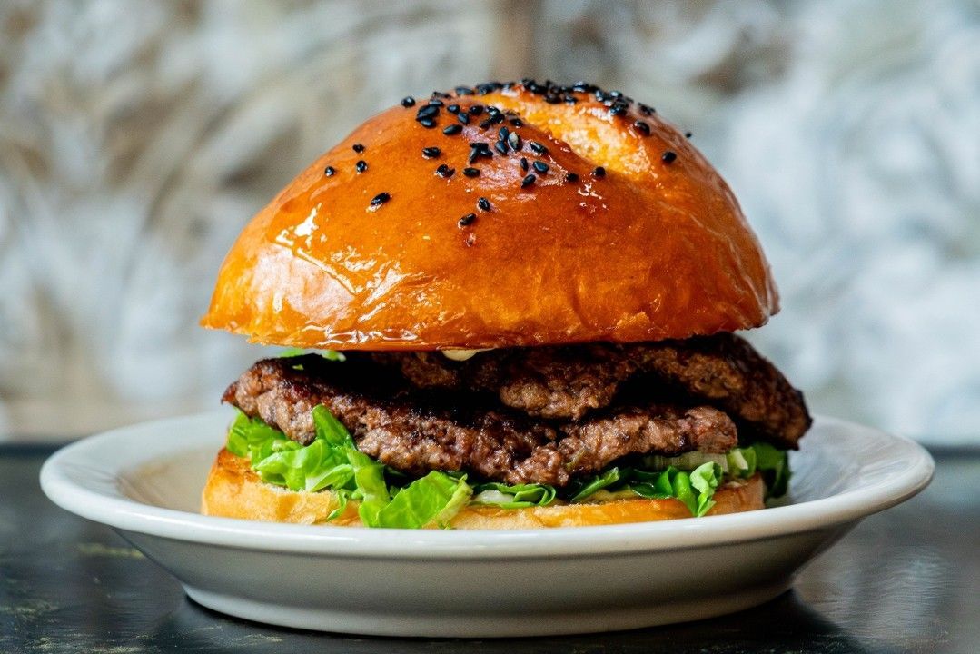 A double-stacked Yuzu Smash Burger from Tiger’s Den, featuring crispy beef patties, fresh lettuce, and a glossy brioche bun sprinkled with black sesame seeds.
