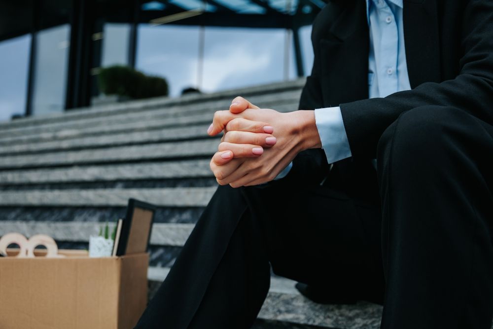 A Person In A Business Suit Sits On Outdoor Stairs With Their Hands Clasped, Beside A Box Of Personal Belongings — Australian Employee Lawyers in Broadbeach Waters, QLD