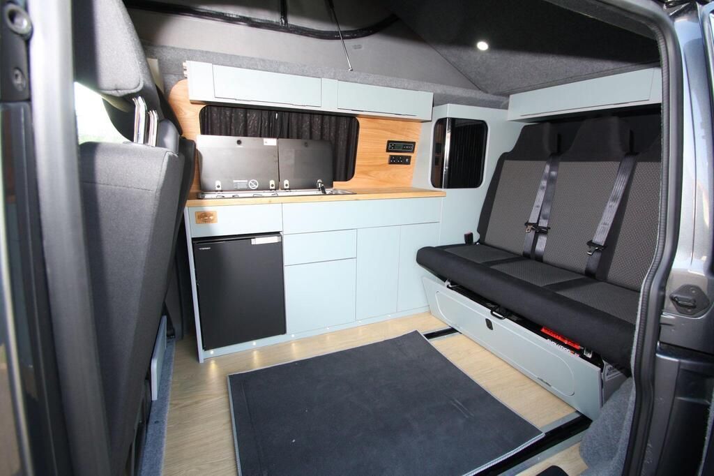 Interior of a compact camper van with a kitchenette, bench seats, and gray furnishings