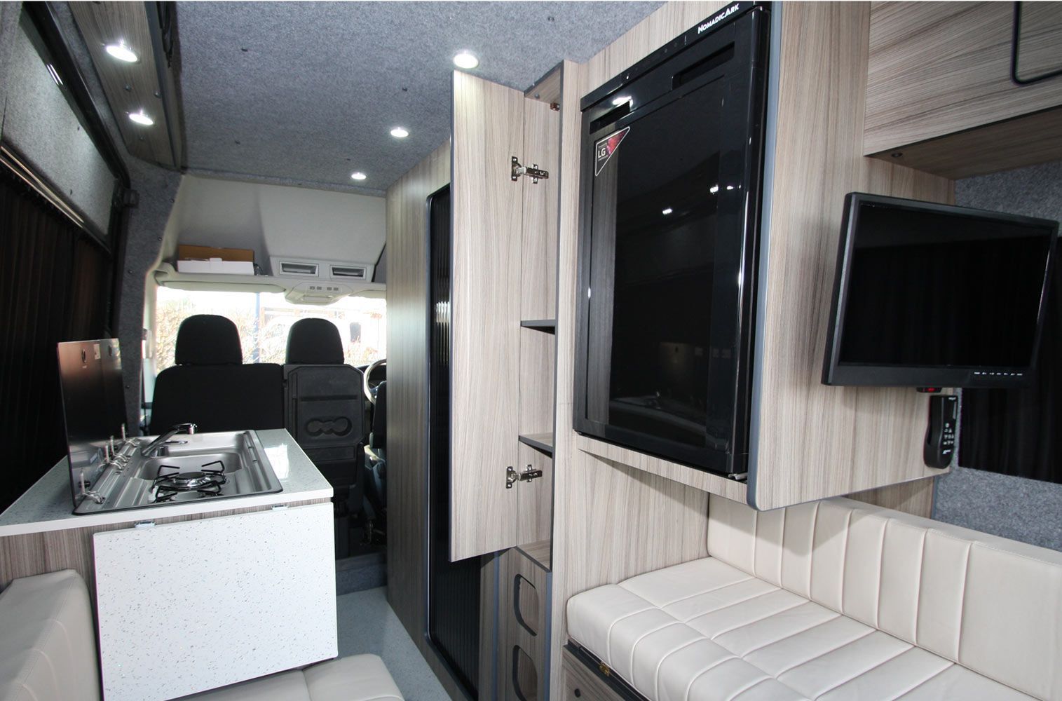 Interior of a modern camper van with white seating, wood cabinets, and a wall-mounted TV.
