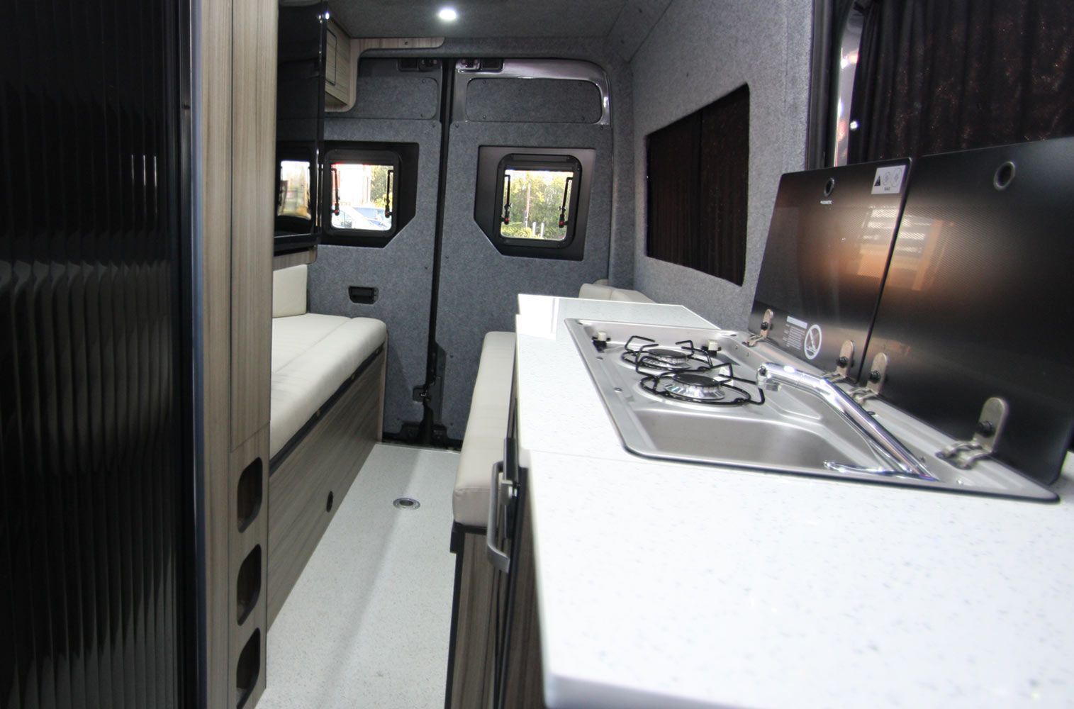 Modern compact kitchen in a narrow RV or tiny home, with white counters, stove, and built-in seating.