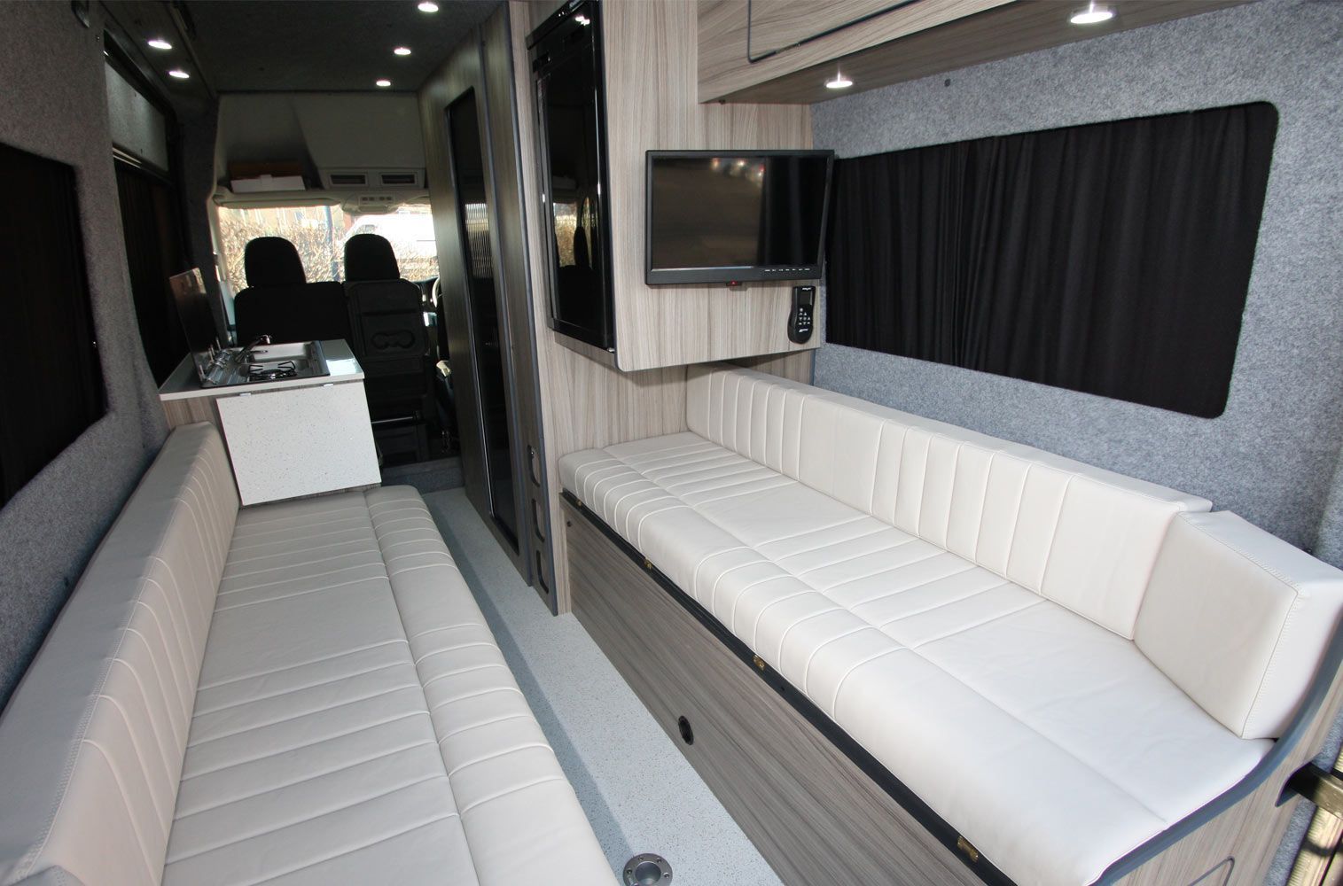 Narrow lounge interior with white cushioned benches, black curtains, and a TV on the wall