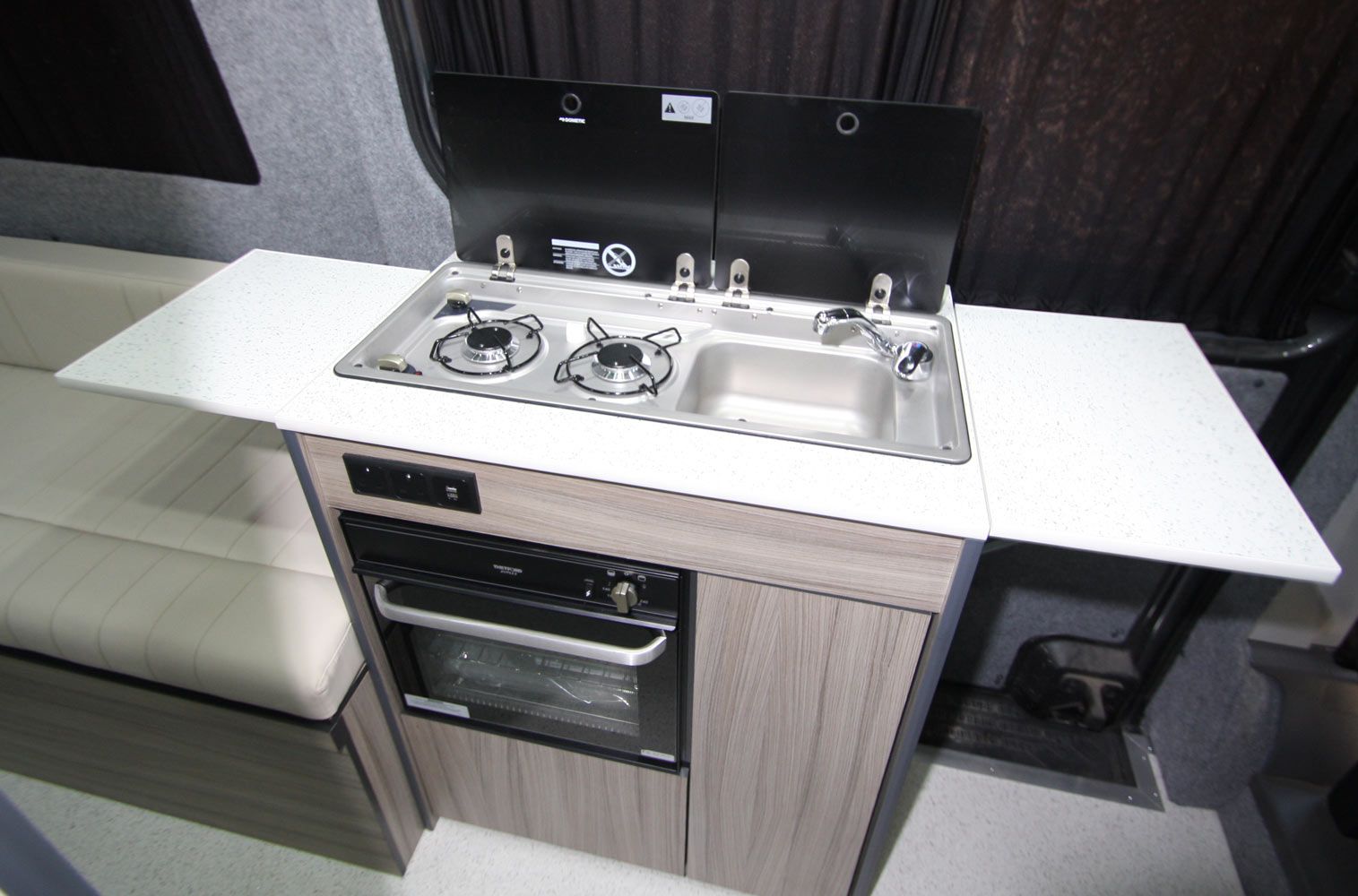 Compact kitchenette with stainless steel sink, two-burner stove, oven, and white countertop.