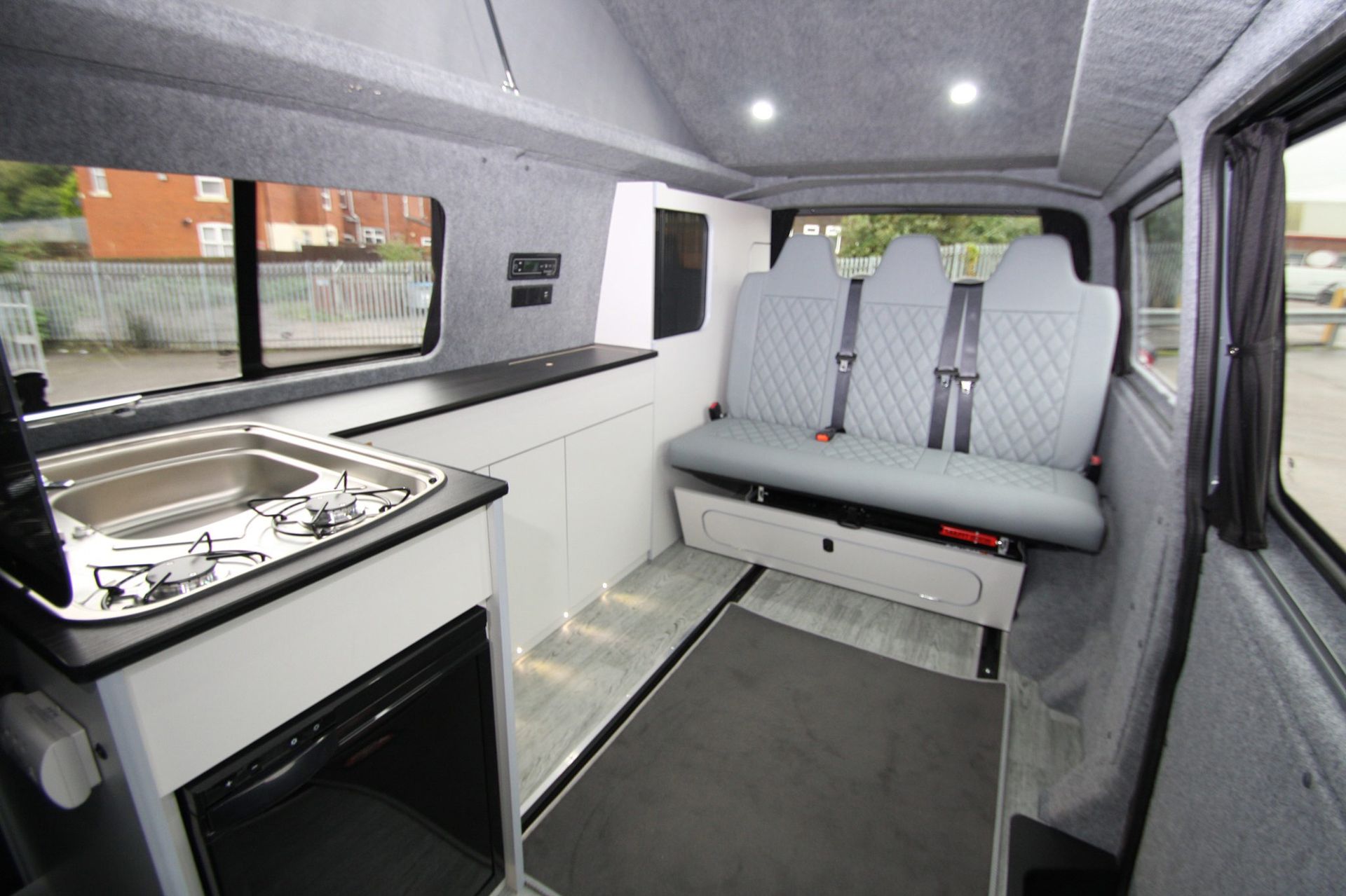 Modern camper van interior with gray seats, white cabinets, stove, and large windows