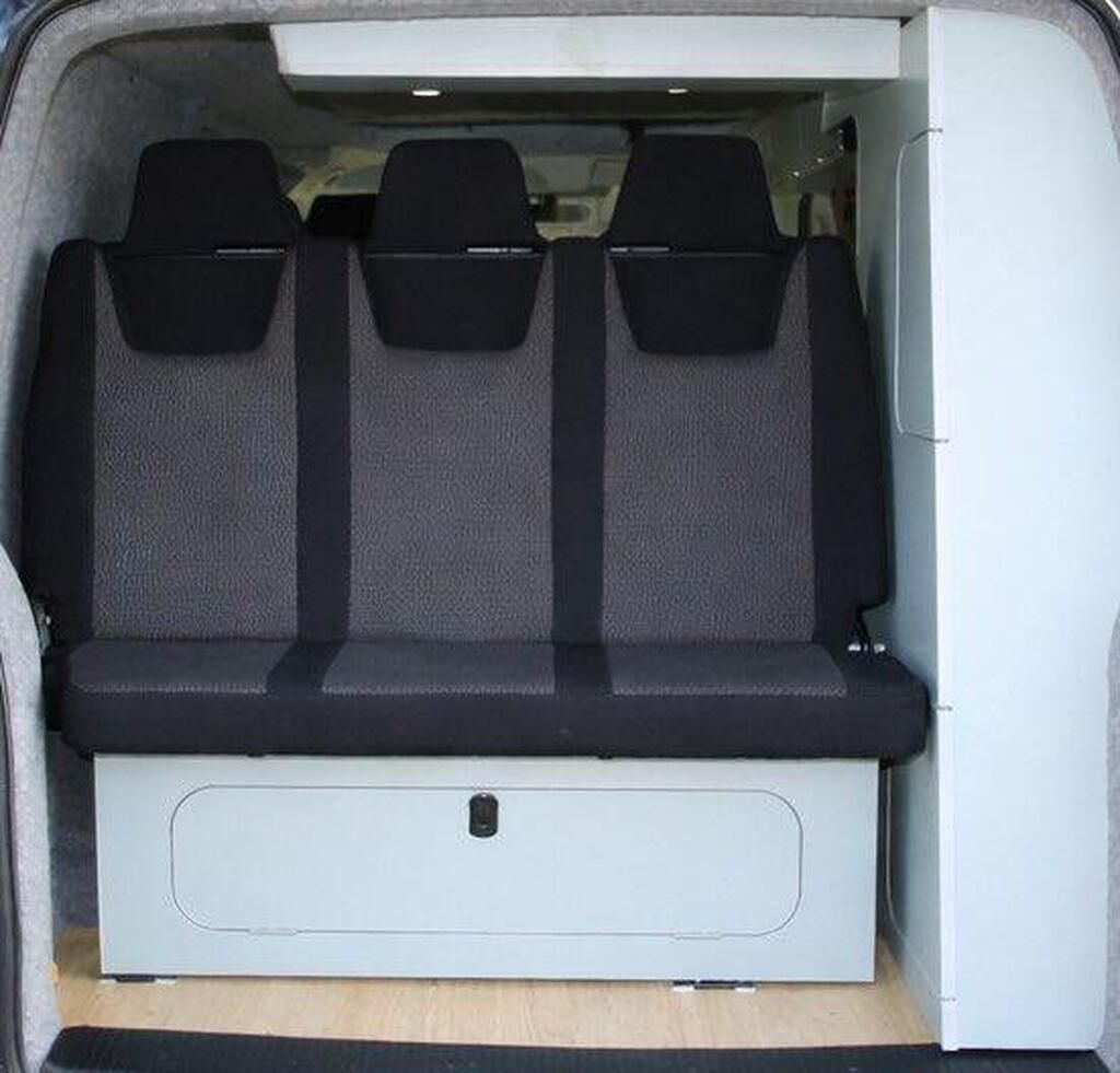Rear van seating area with three black-and-gray seats and a white storage panel below.