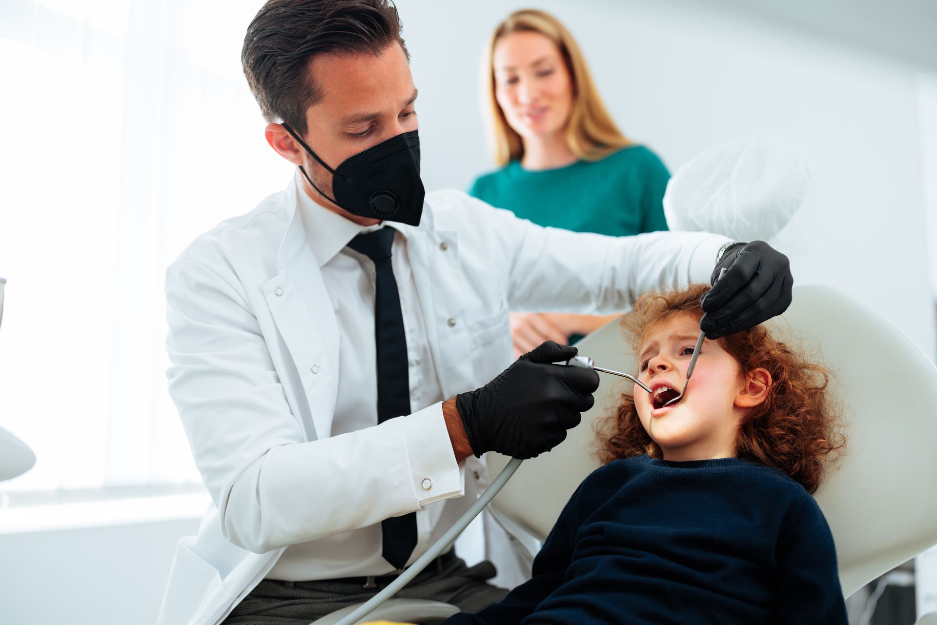 A dentist wearing a mask examines a young child's teeth with dental tools while a parent watches