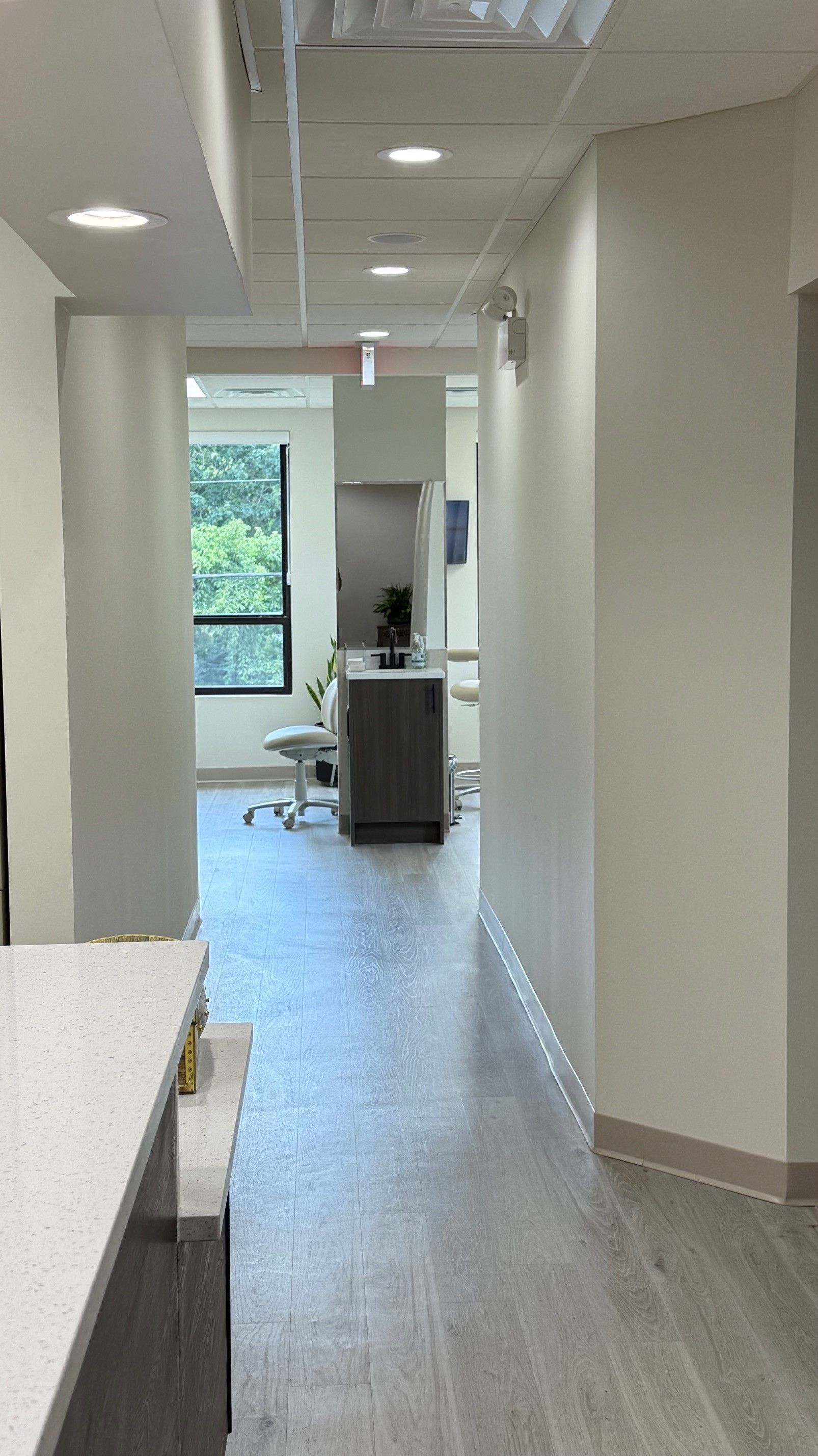 Narrow office hallway with light gray walls, dark wood-look floor, and view into treatment rooms.