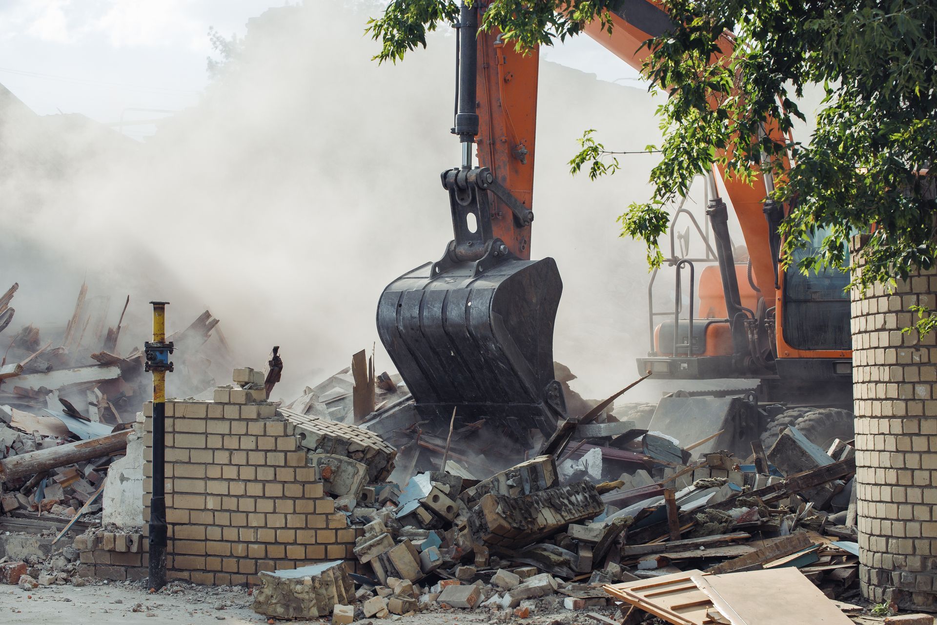 Orange excavator demolishes a brick building, creating a cloud of dust.