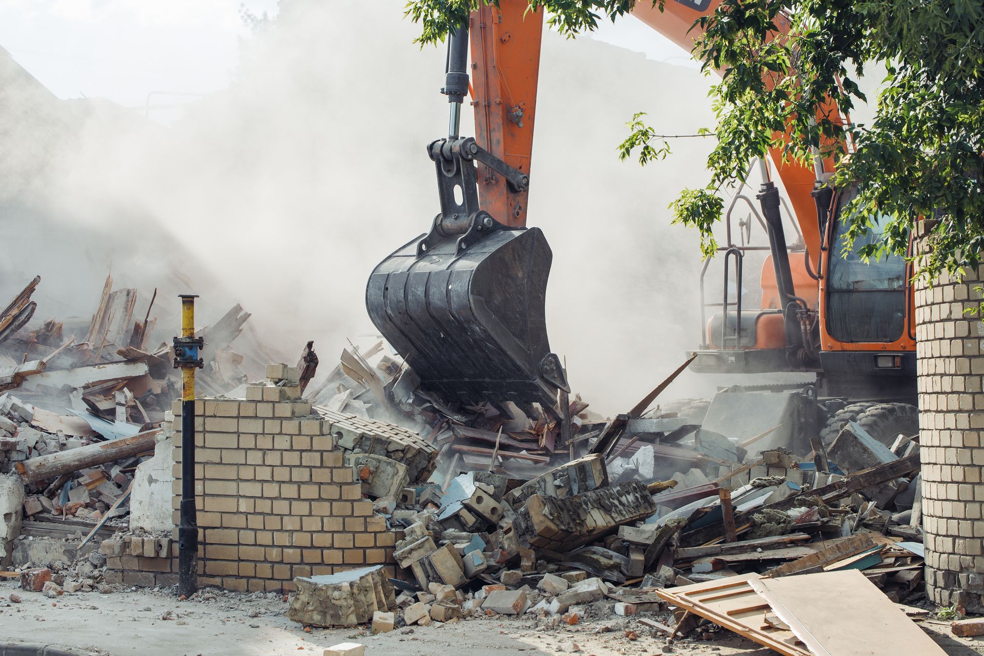 A small excavator demolishes a concrete wall next to a building; debris is scattered.