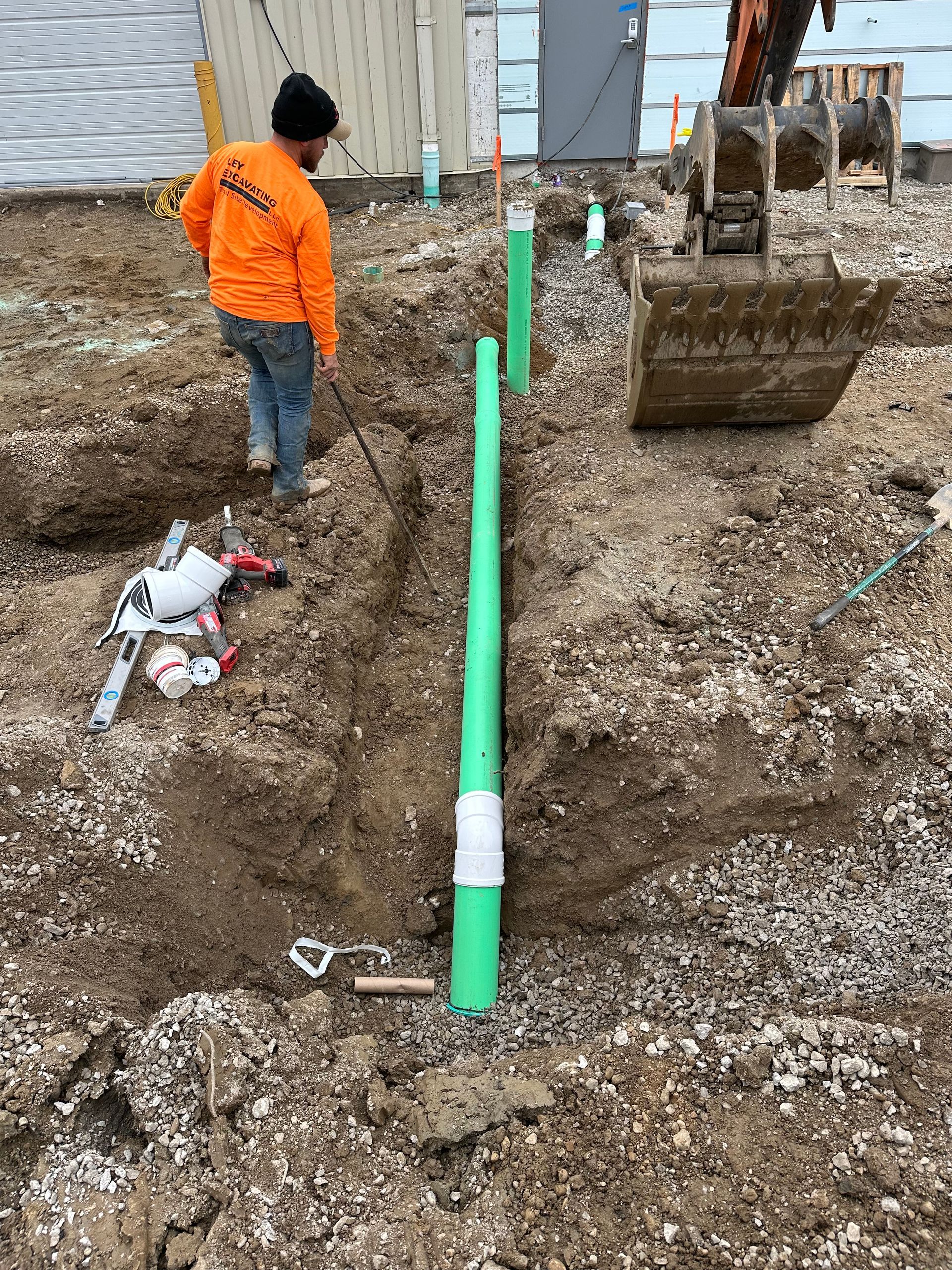 Trench dug in dirt with two black pipes laid inside, beside a concrete sidewalk and green grass.