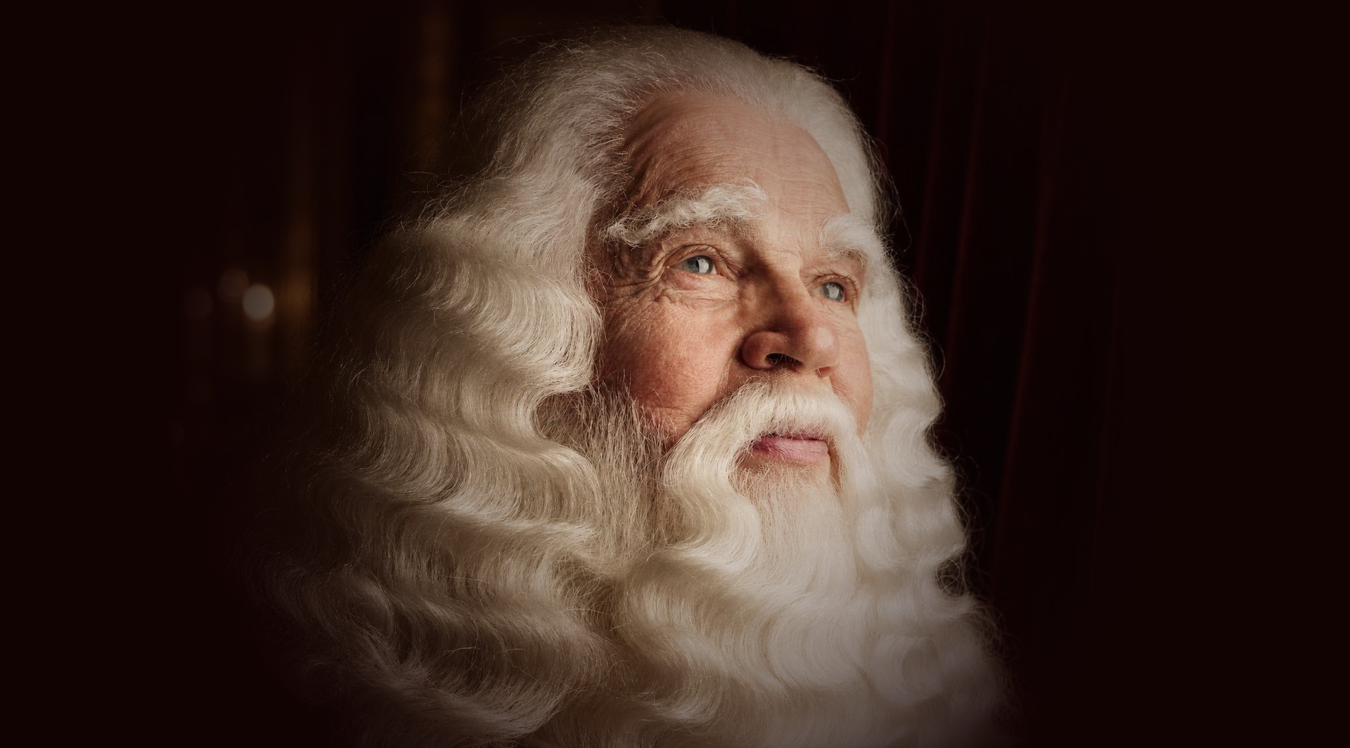 Portret van een oudere man met een witte baard en lang, krullend wit haar tegen een donkere achtergrond.