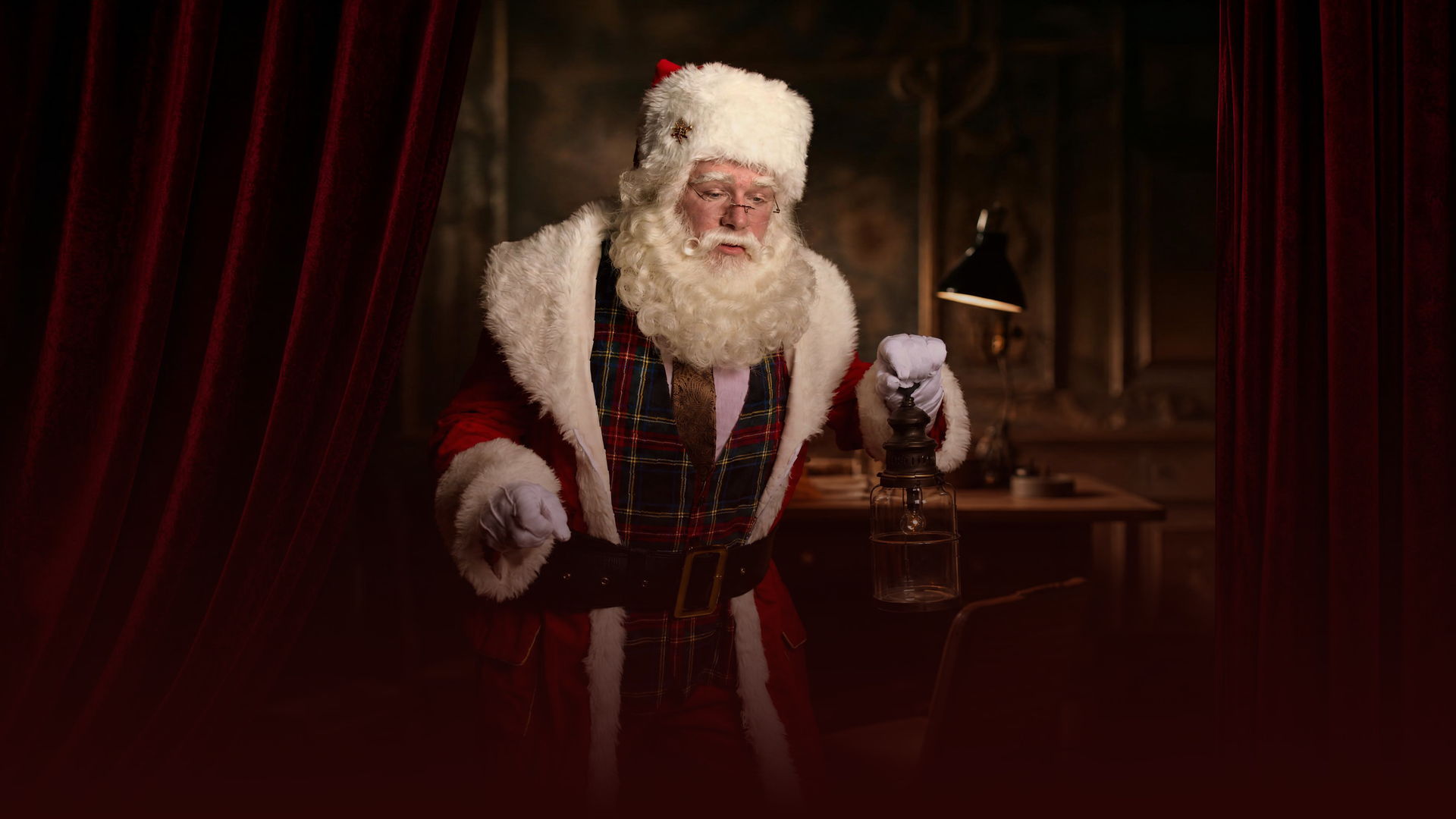 De Kerstman in een rood pak staat in een schemerige kamer naast een klein brandend lampje en een bureau.