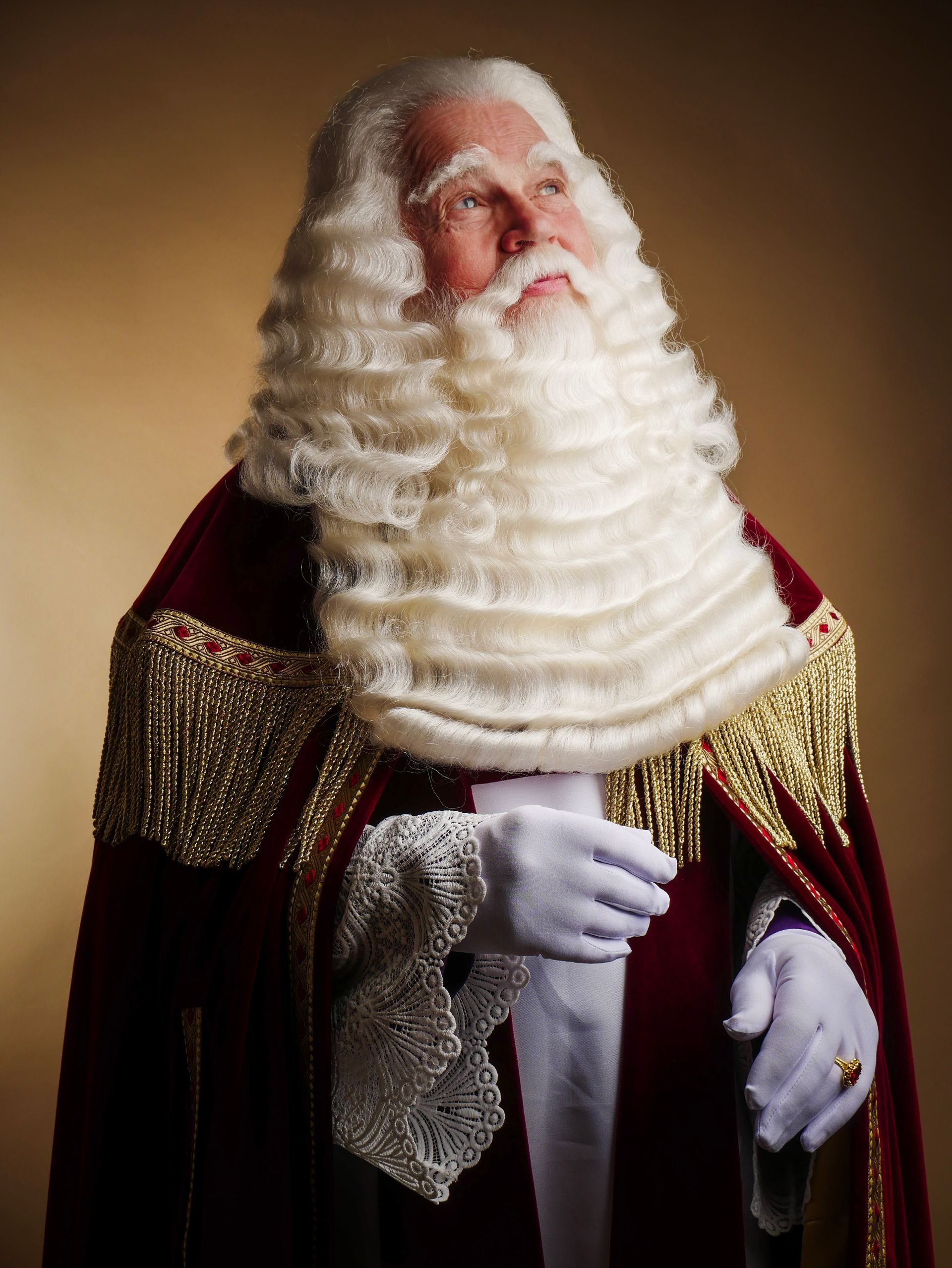 sint in een rood fluwelen pak met gouden versieringen en een lange witte baard, staand tegen een warme bruine achtergrond.