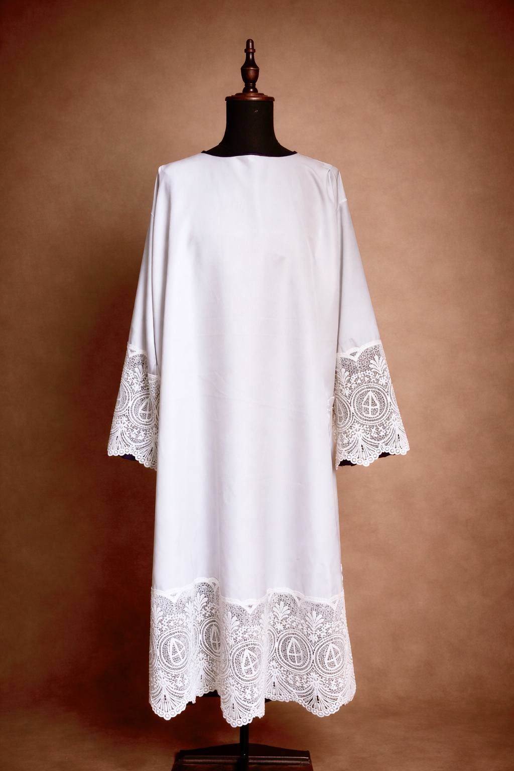 Witte albe met kanten details, getoond op een mannequin tegen een bruine achtergrond.