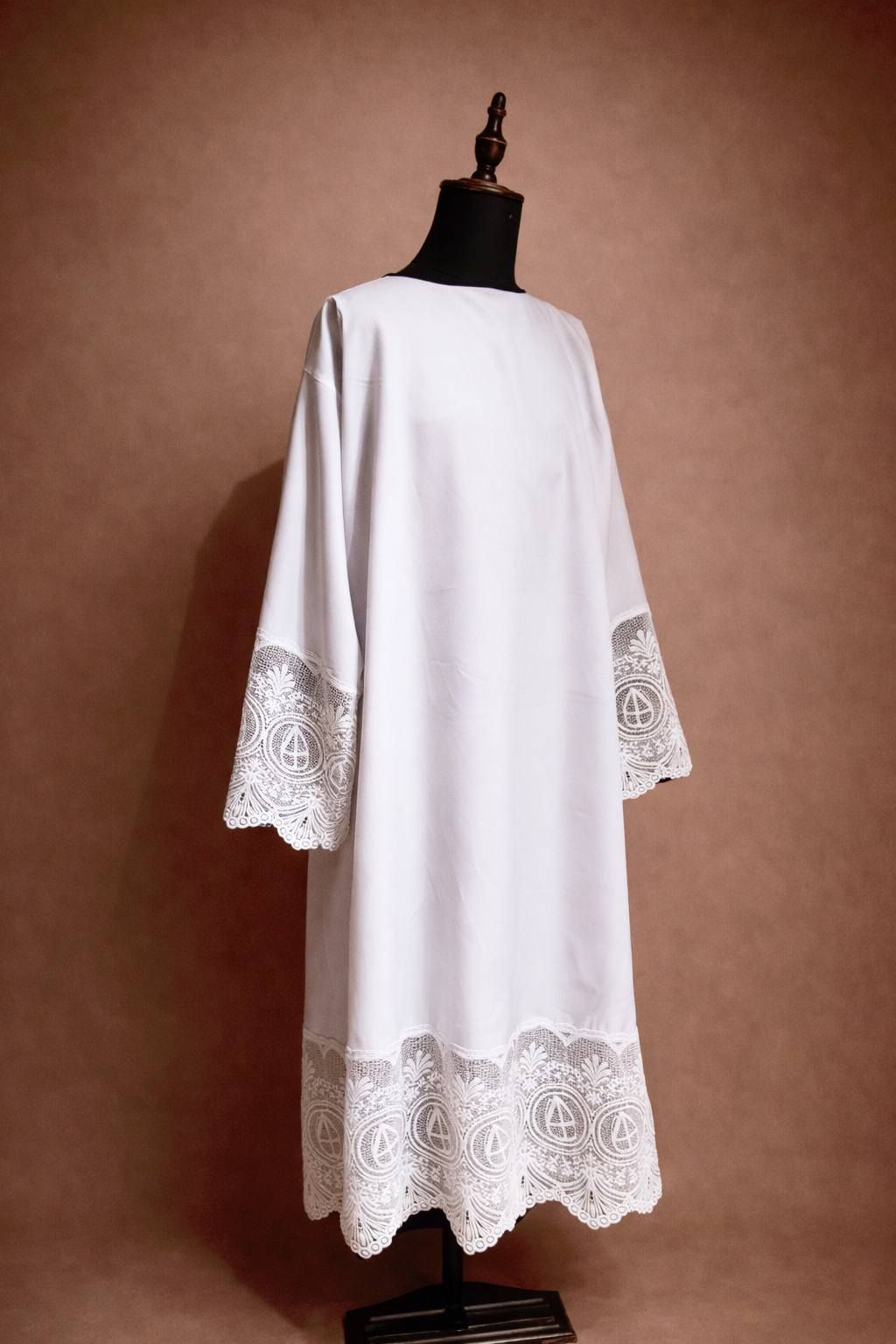 Witte albe met kanten details, getoond op een mannequin tegen een bruine achtergrond.