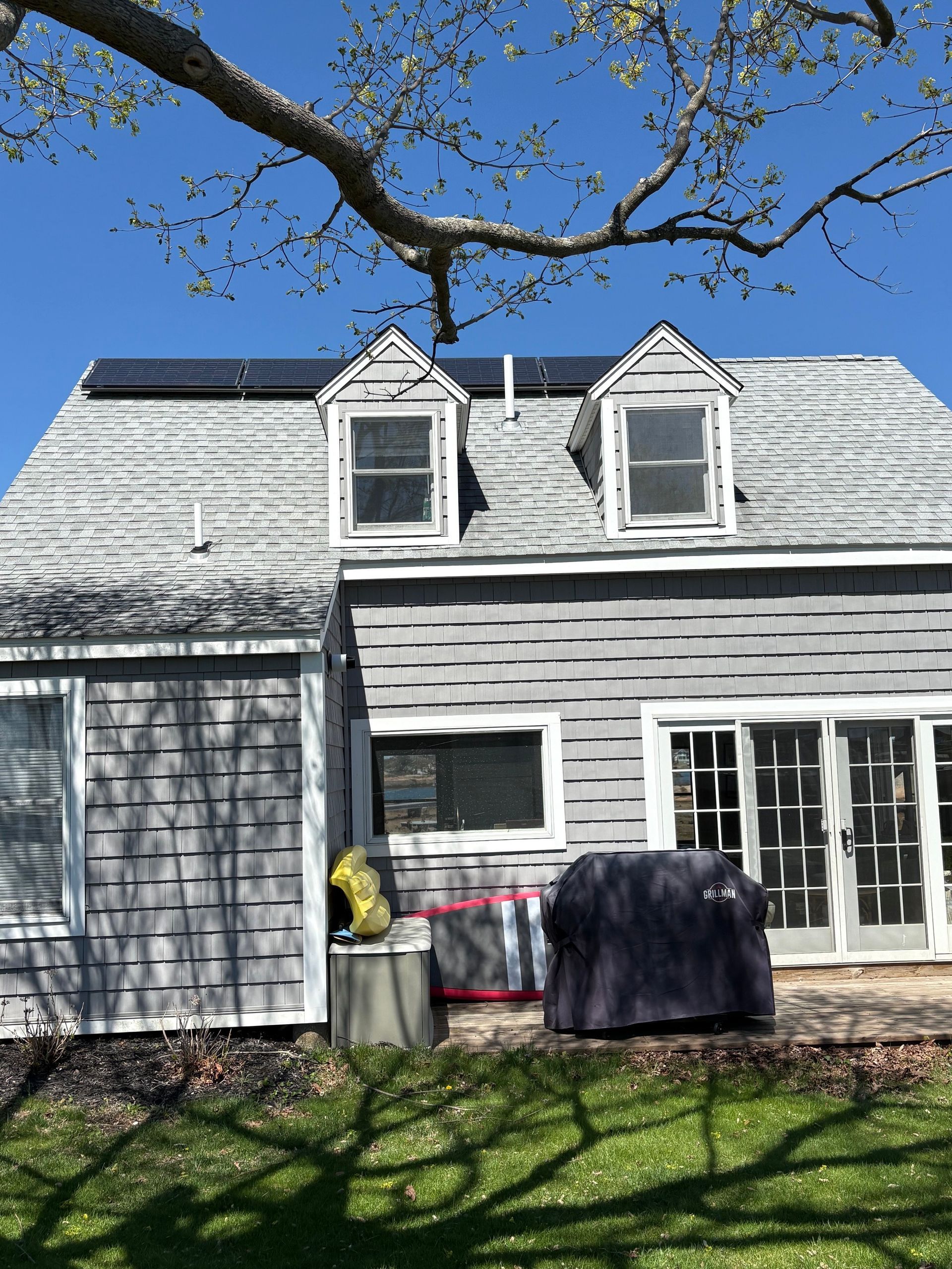 Gray house with dormer windows, white-trimmed doors, and a bare tree branch overhead on a sunny day