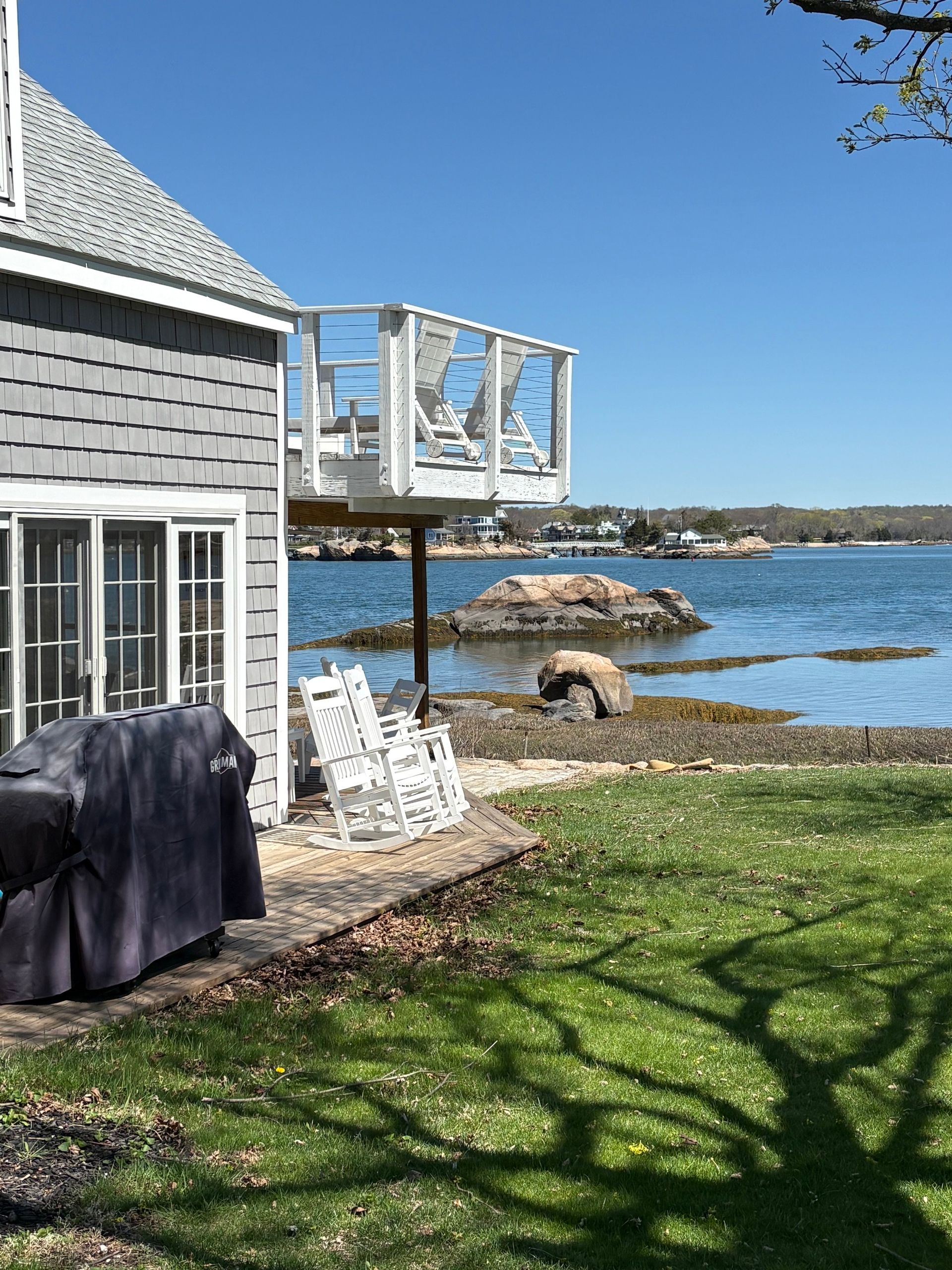 Coastal house with white deck overlooking calm blue water, green lawn, and rocky shoreline under clear sky