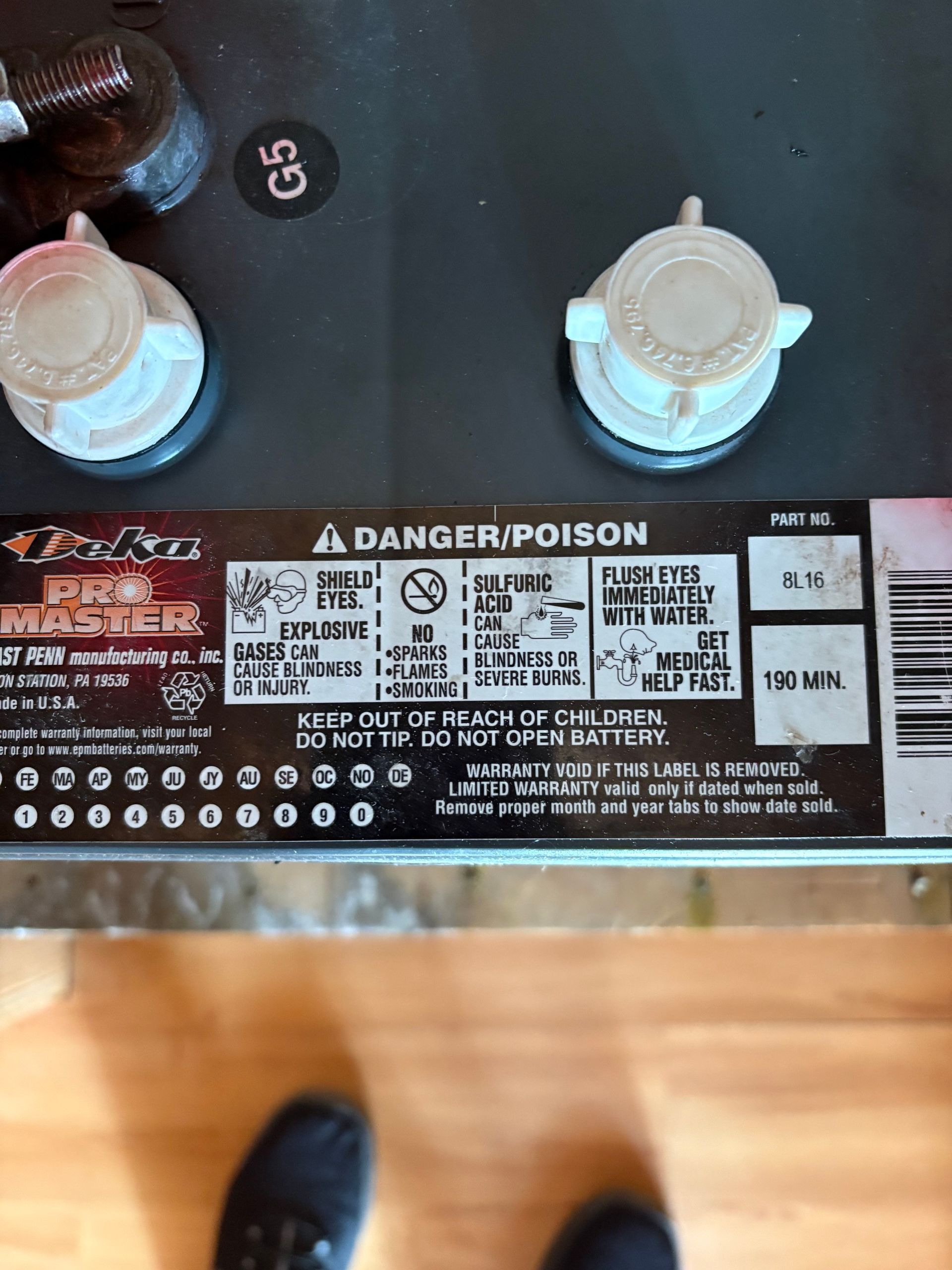 Close-up of a car battery top with warning labels, terminals, and fluid caps on a garage floor.