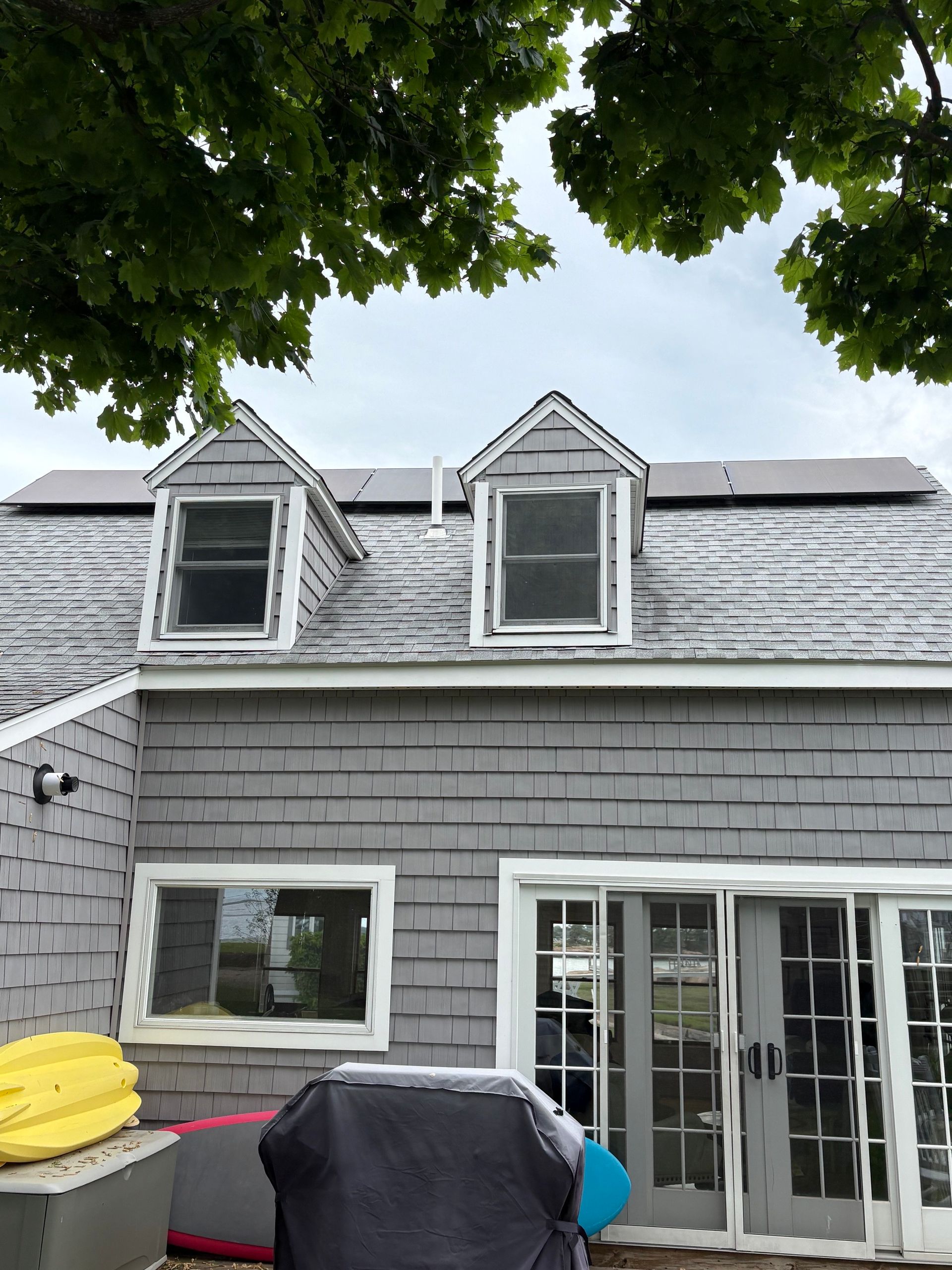 Gray house exterior with two dormer windows, patio doors, and trees overhead