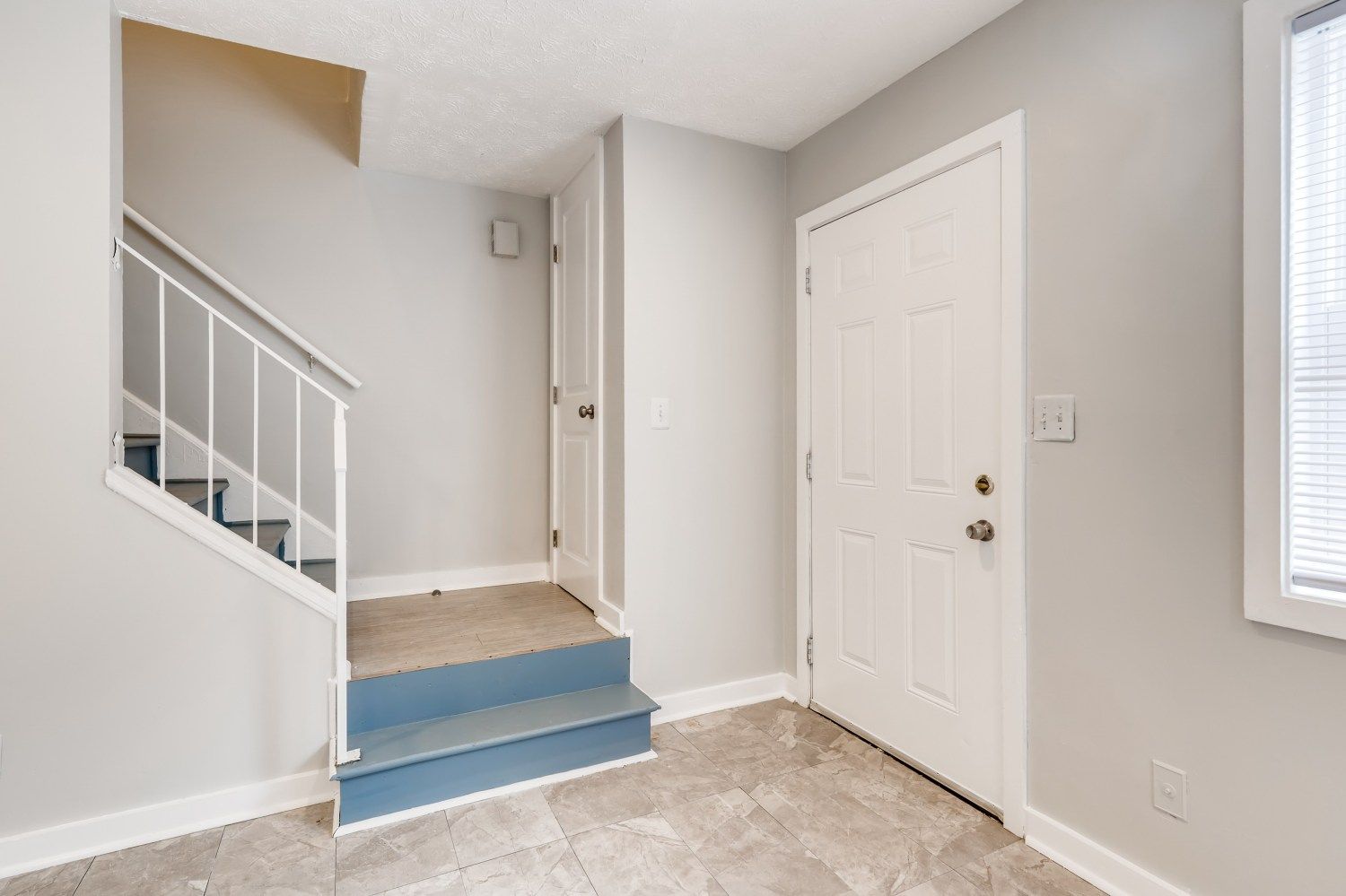 Bright entryway with white door, blue carpeted steps, and staircase railing against gray walls.