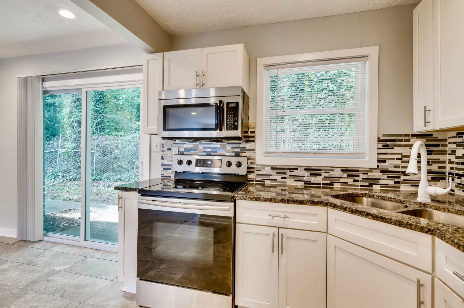 Bright kitchen with white cabinets, stainless steel oven, granite counters, and sliding glass door to a wooded view
